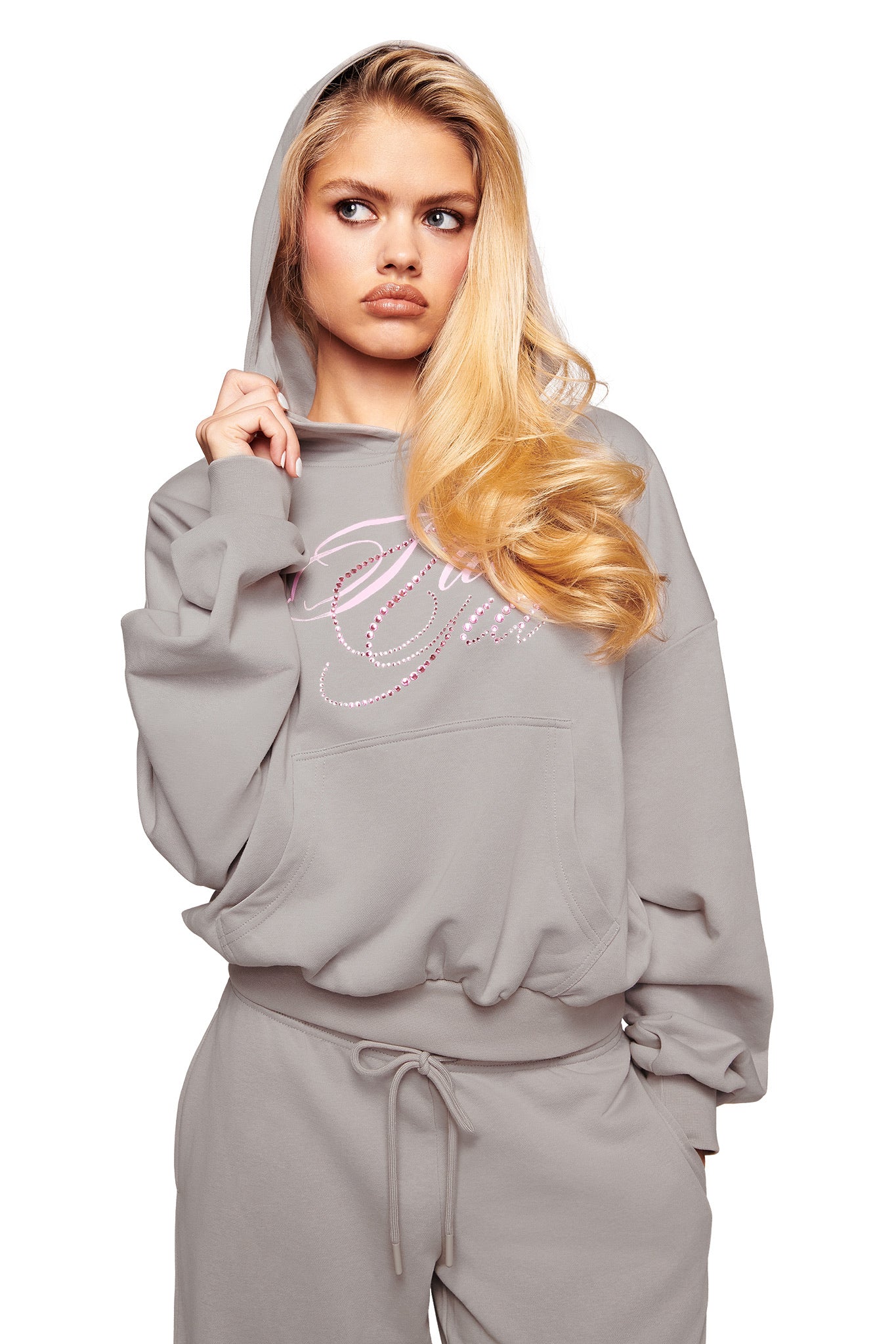 A young woman with long blonde hair models the I.AM.GIA KADE HOODIE in grey, paired with matching sweatpants. She stands against a white background, holding the hood with one hand and gazing seriously to the side.