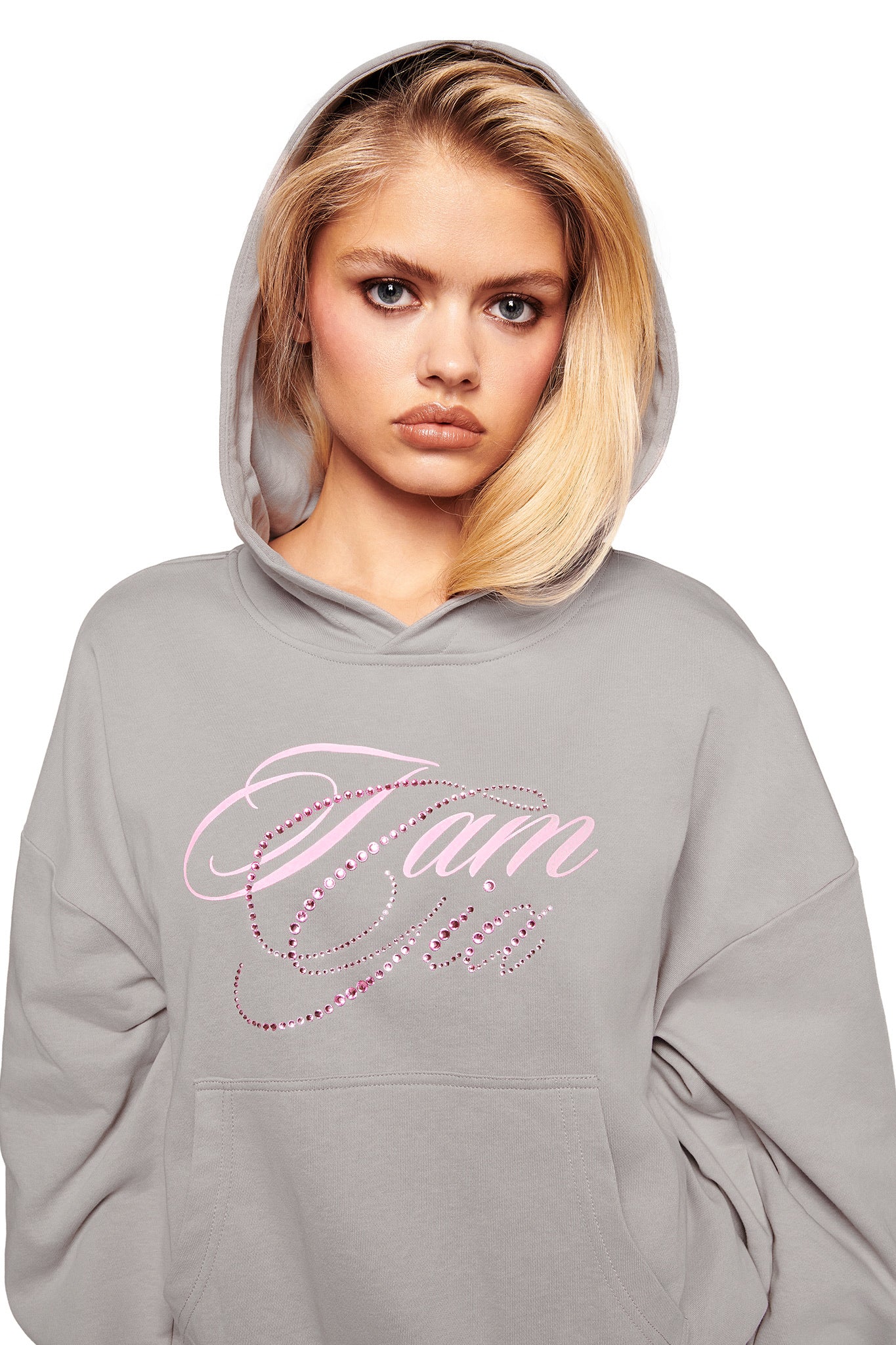 A person with long blonde hair wears the I.AM.GIA KADE HOODIE in grey, hood up. "I am Gia" is scripted in pink and white on the front as they face the camera with a neutral expression.