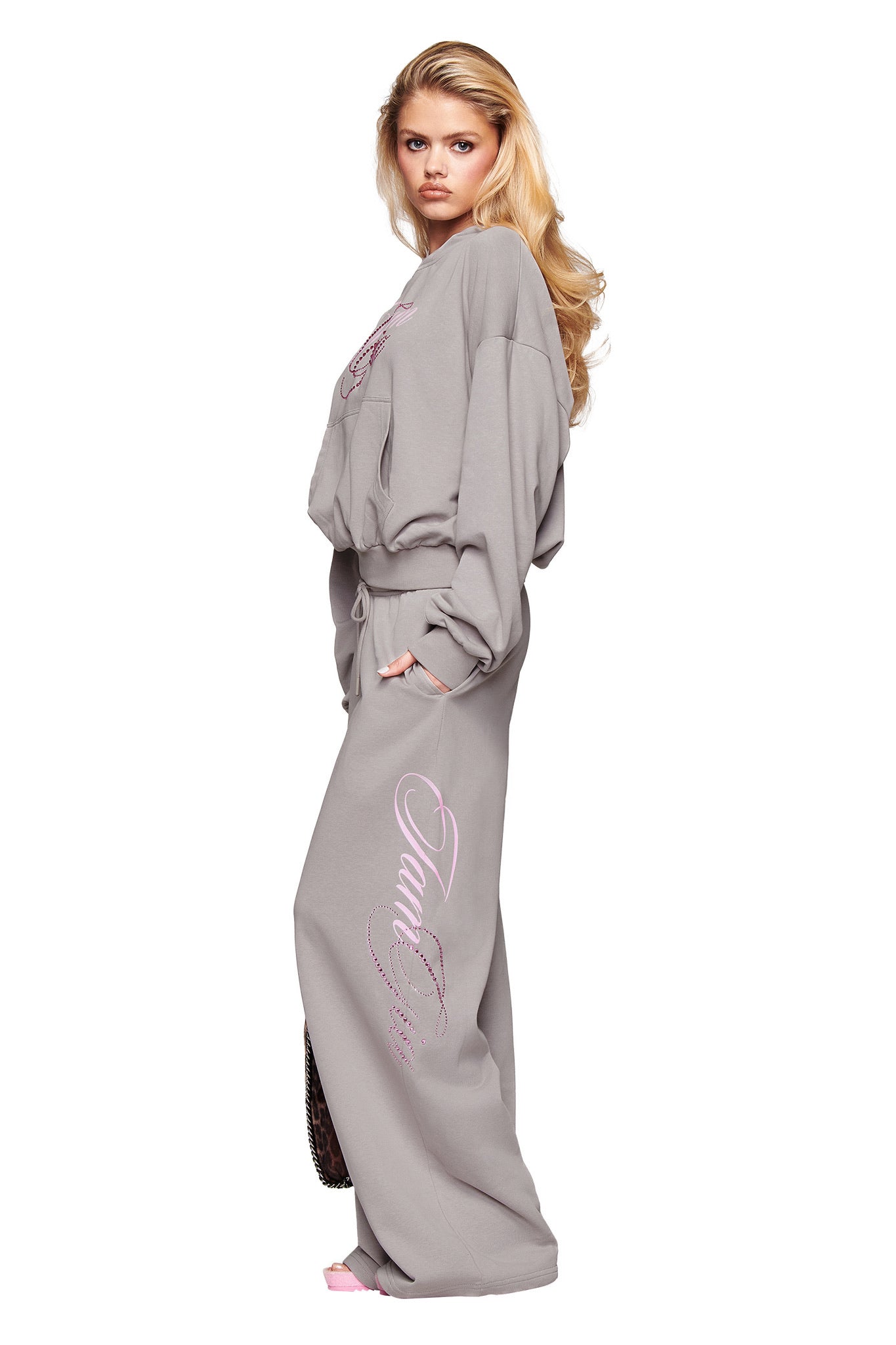 A woman with long blonde hair models the I.AM.GIA KADE PANT in grey, featuring pink script along the side. She stands in profile against a white background, highlighting the pants’ relaxed, wide-leg fit.