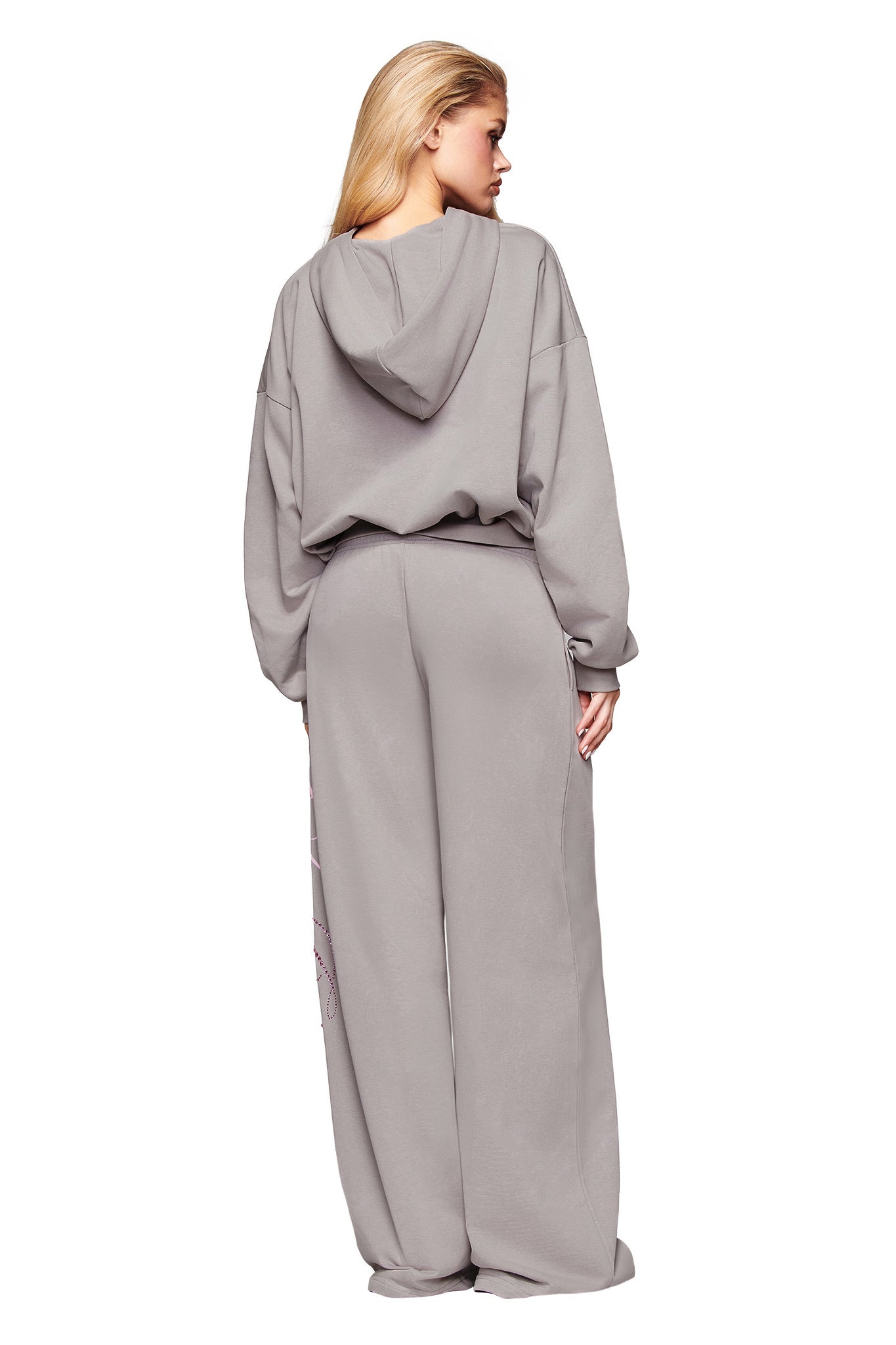 A woman with blonde hair down faces away, wearing the I.AM.GIA KADE HOODIE in grey and matching wide-leg sweatpants. Her right hand is on her left hip against a white background.