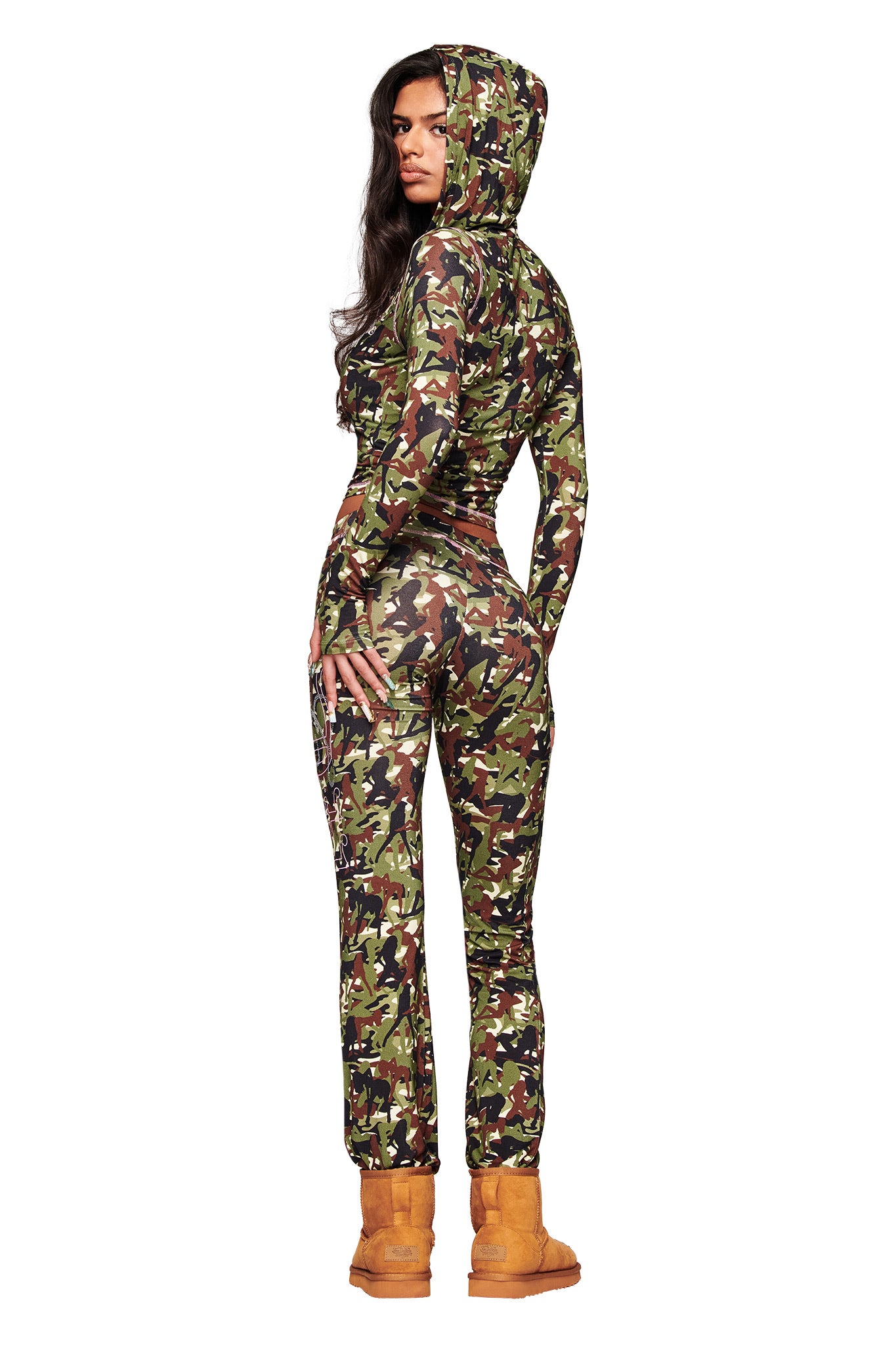 A woman faces backward in the I.AM.GIA X VON DUTCH BLARE TRACKPANT - GIA GIRL CAMO by I.AM.GIA, paired with tan boots. Her long dark hair falls as she glances over her shoulder, channeling Von Dutch style against a white background.