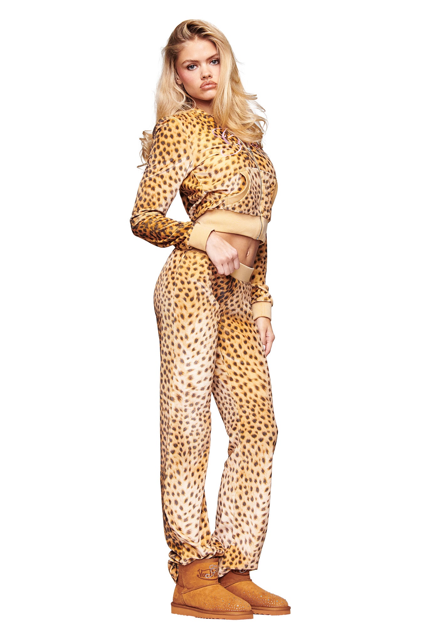 A woman with long blonde hair poses confidently in the I.AM.GIA X VON DUTCH Kameron Velour Trackpant set in leopard print, featuring a matching cropped zip-up hoodie and pants, styled with tan boots against a white background.