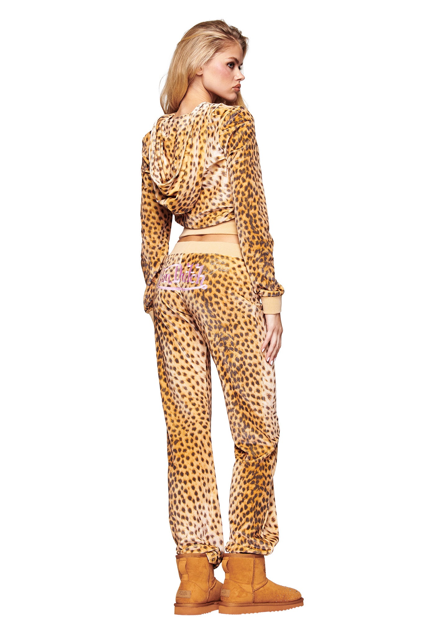 A woman with long blonde hair and tan suede boots poses facing away in the I.AM.GIA X VON DUTCH KAMERON VELOUR JACKET - LEOPARD and matching pants, featuring a pink graphic on the back.
