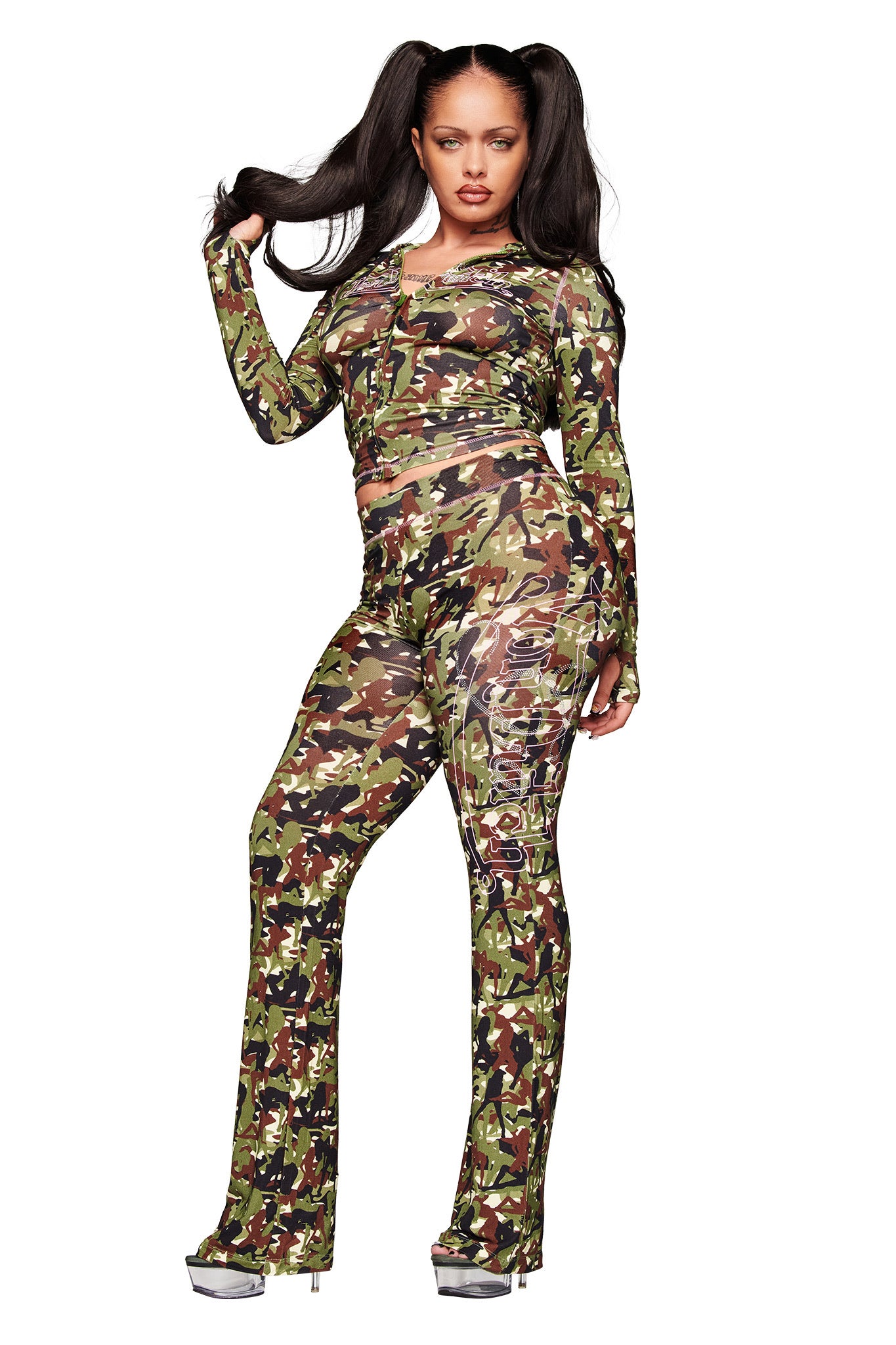 A woman with long dark pigtails models the I.AM.GIA X VON DUTCH BLARE TRACKPANT - GIA GIRL CAMO and a matching sheer top, paired with high-heeled sandals, against a white background.