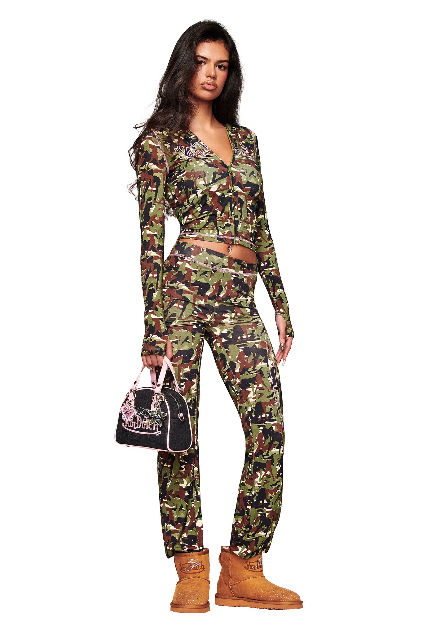 A woman exudes confidence in the I.AM.GIA X Von Dutch Blare Hoodie - Gia Girl Camo, paired with matching pants, tan boots, and a black Juicy Couture handbag with pink handles for a bold, stylish look.