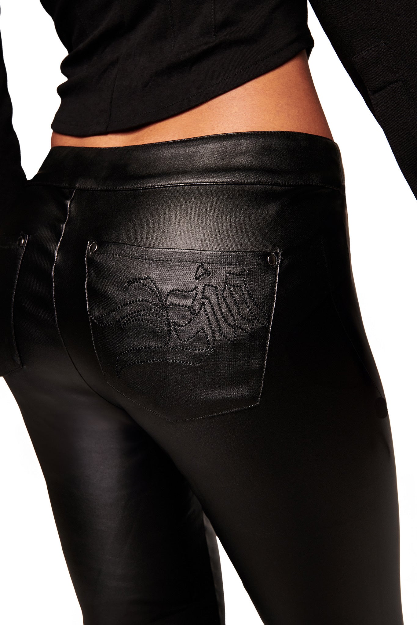 A close-up of a person wearing the I.AM.GIA JETT PANT - BLACK, featuring shiny black skinny jeans with a waxed finish and embroidered back pocket detail, paired with a black jacket against a plain white background.