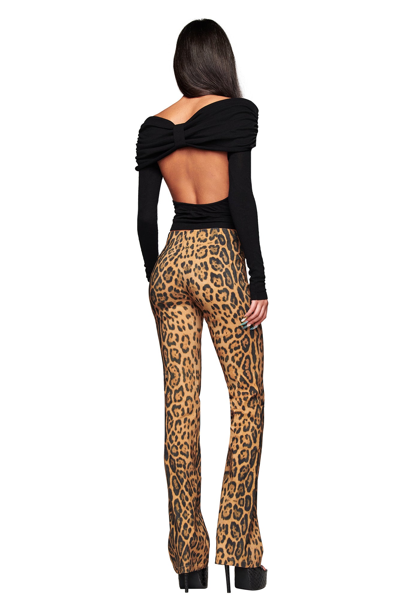 A woman faces away, wearing a black long-sleeve backless top with a bow and I.AM.GIA UMA PANT - PANTHERA high-waisted, flared animal print pants, paired with black platform heels.