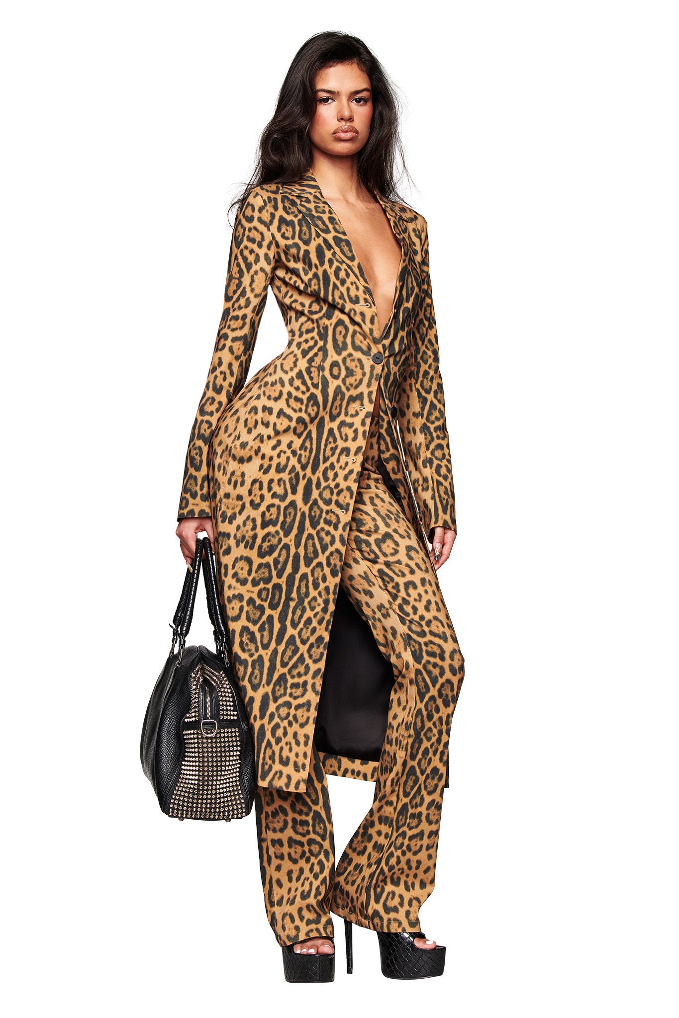 A woman with long dark hair and a serious expression stands confidently against a white background, wearing I.AM.GIA’s UMA PANT - PANTHERA flared pants with a matching animal print coat, black studded handbag, and black platform heels.