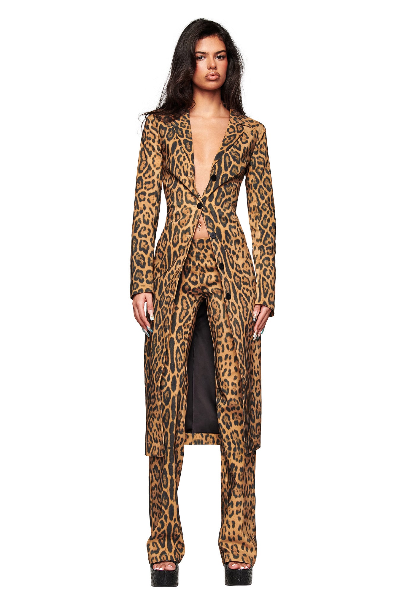 A person stands facing forward in I.AM.GIA’s UMA PANT - PANTHERA, featuring a long, fitted leopard-print coat with matching Panthera print pants. The buttoned coat has a deep V neckline and is styled with black platform shoes.