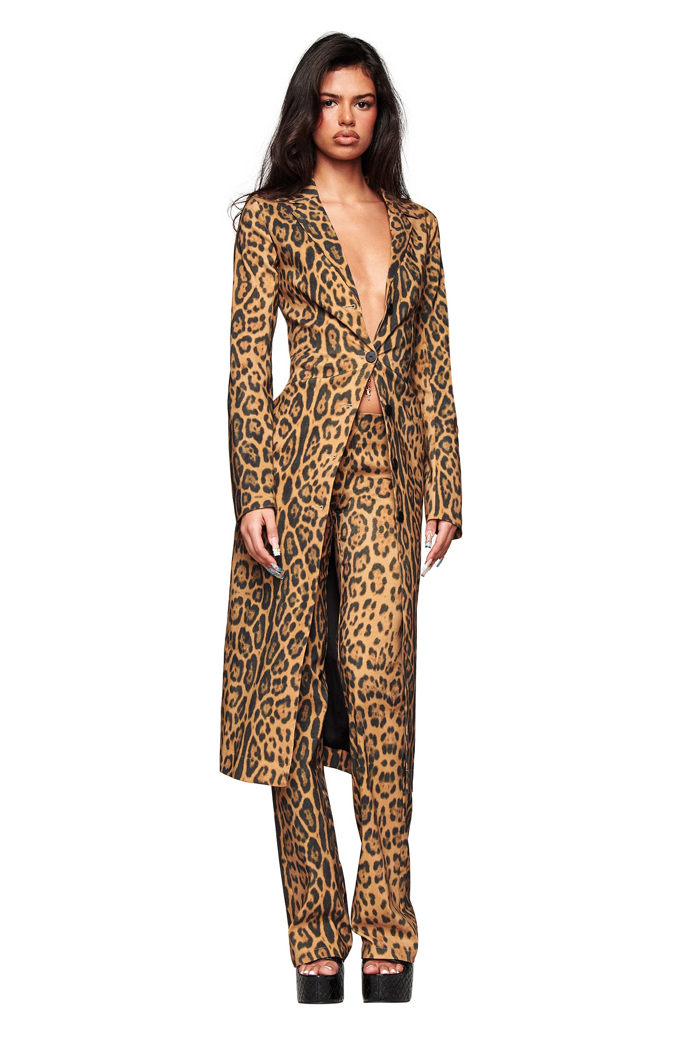A woman with long dark hair faces forward in I.AM.GIA's UMA PANT - PANTHERA, featuring flared Panthera animal print. She pairs it with a matching open-front coat and black platform shoes, posing against a white background.