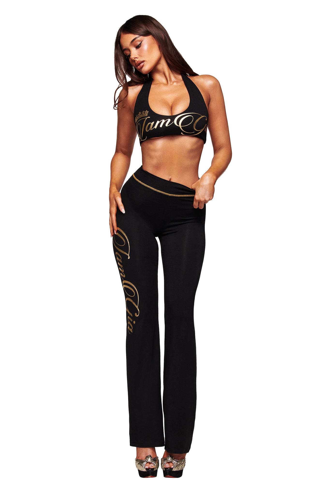 A woman poses against a white background in the I.AM.GIA BLARE FORM TRACKPANT in black and gold, styled with a matching black halter crop top and high-heeled shoes. Both pieces have a body-hugging fit and gold script text.