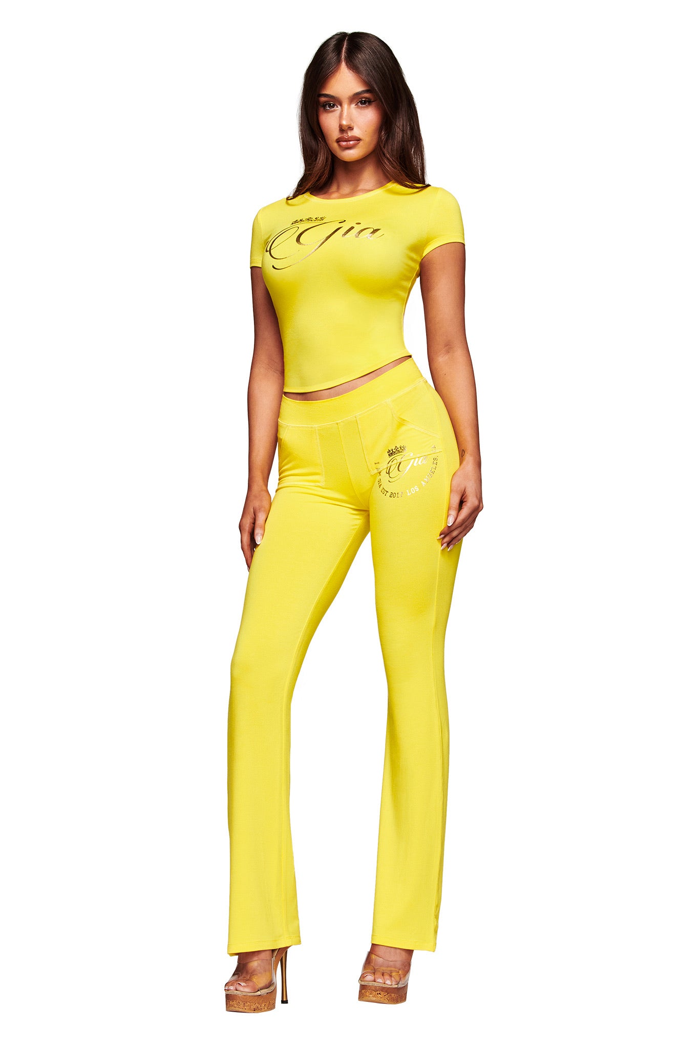 A woman with long brown hair faces forward, wearing the I.AM.GIA BLARE FORM TEE - YELLOW GOLD FOIL and matching high-waisted pants with text details, both bodycon fit. She pairs them with heels against a white background.