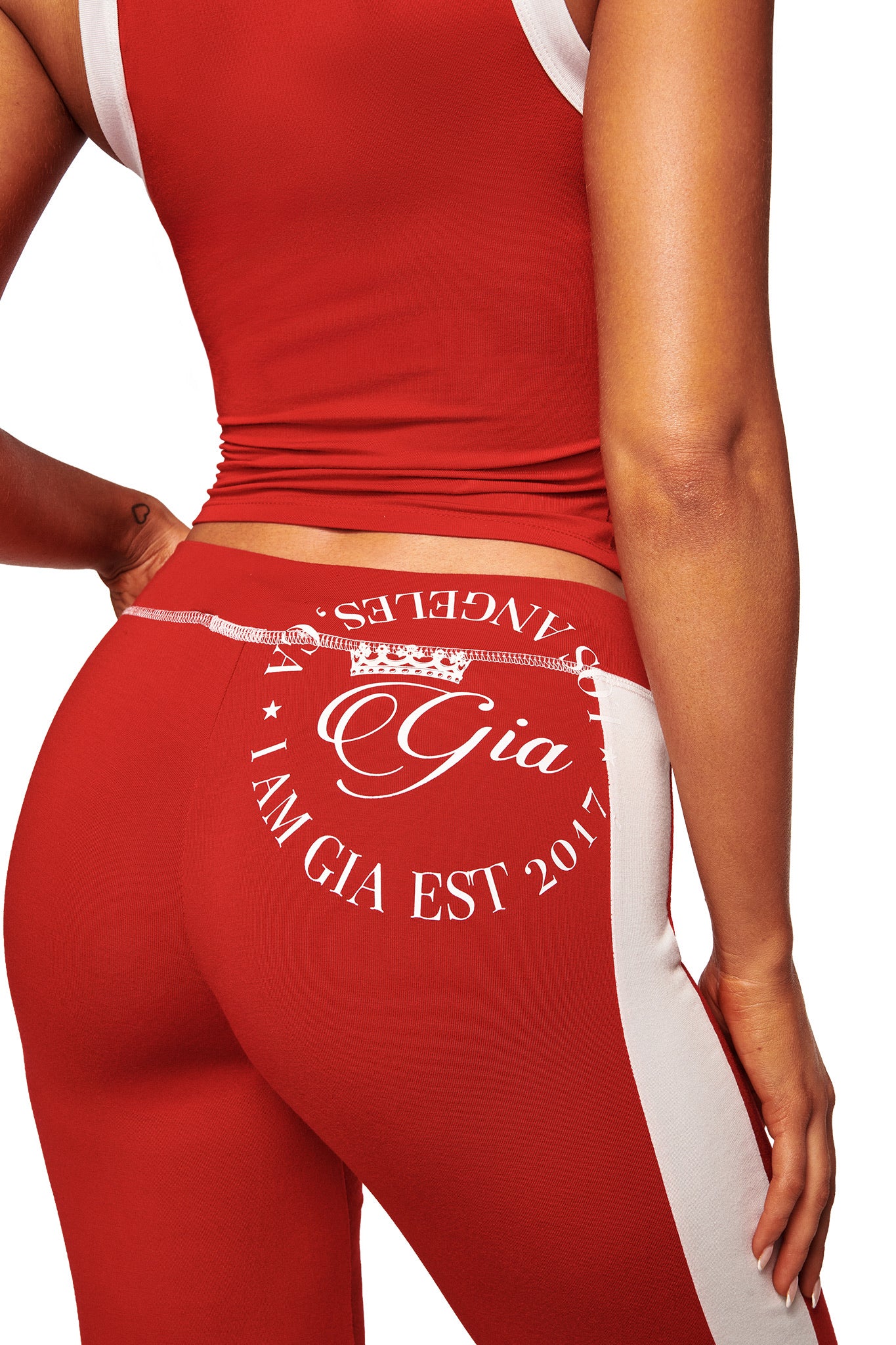 A woman wears the I.AM.GIA BLARE PANELLED TRACKPANT in red, featuring a straight leg and "Gia Los Angeles I AM GIA EST 2017" print on the back, shown in a close-up of her midsection and hip.