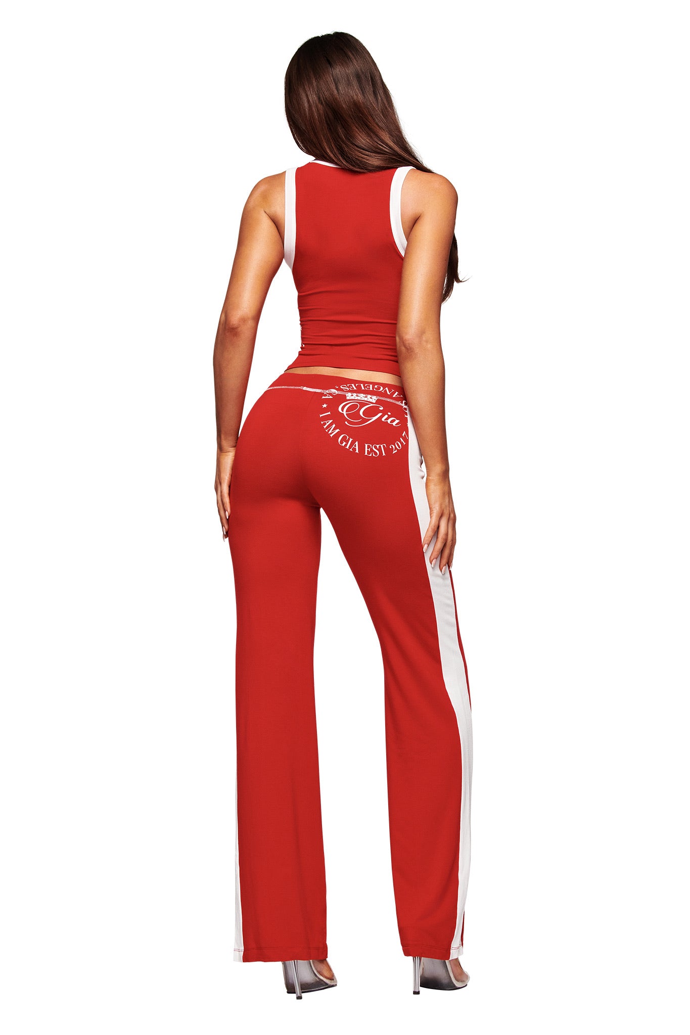 A woman with long brown hair stands with her back to the camera, wearing high heels and the I.AM.GIA BLARE BIND TANK in red, paired with matching wide-leg pants featuring text on the back.