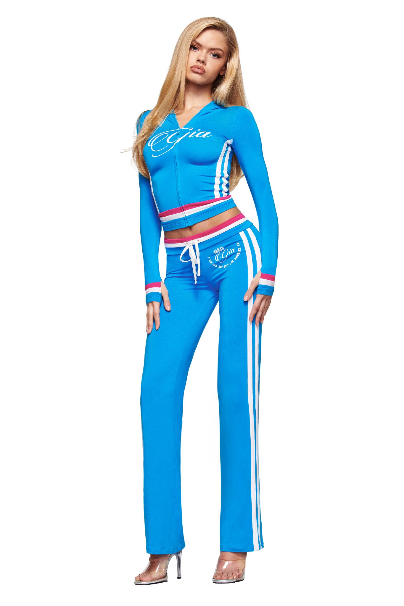 A woman with long blonde hair wears I.AM.GIA's BLARE STRIPED TRACKPANT in sky blue, styled with a matching cropped zip-up hoodie. She poses in clear high heels against a plain white background.