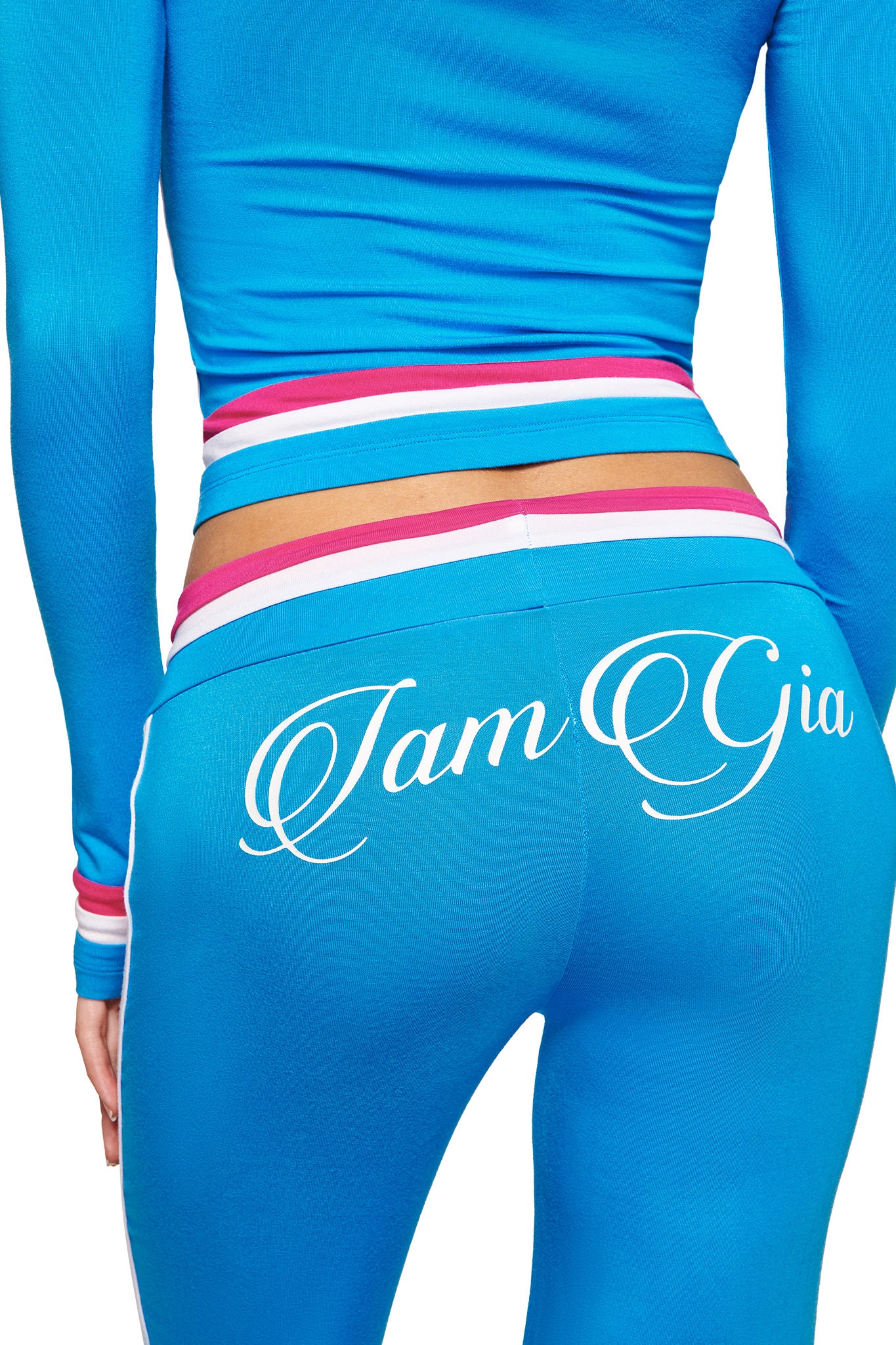 Someone wears the I.AM.GIA BLARE STRIPED TRACKPANT in sky blue with a matching top and colorful striped waistband, featuring “I am Gia” in white script across the back.