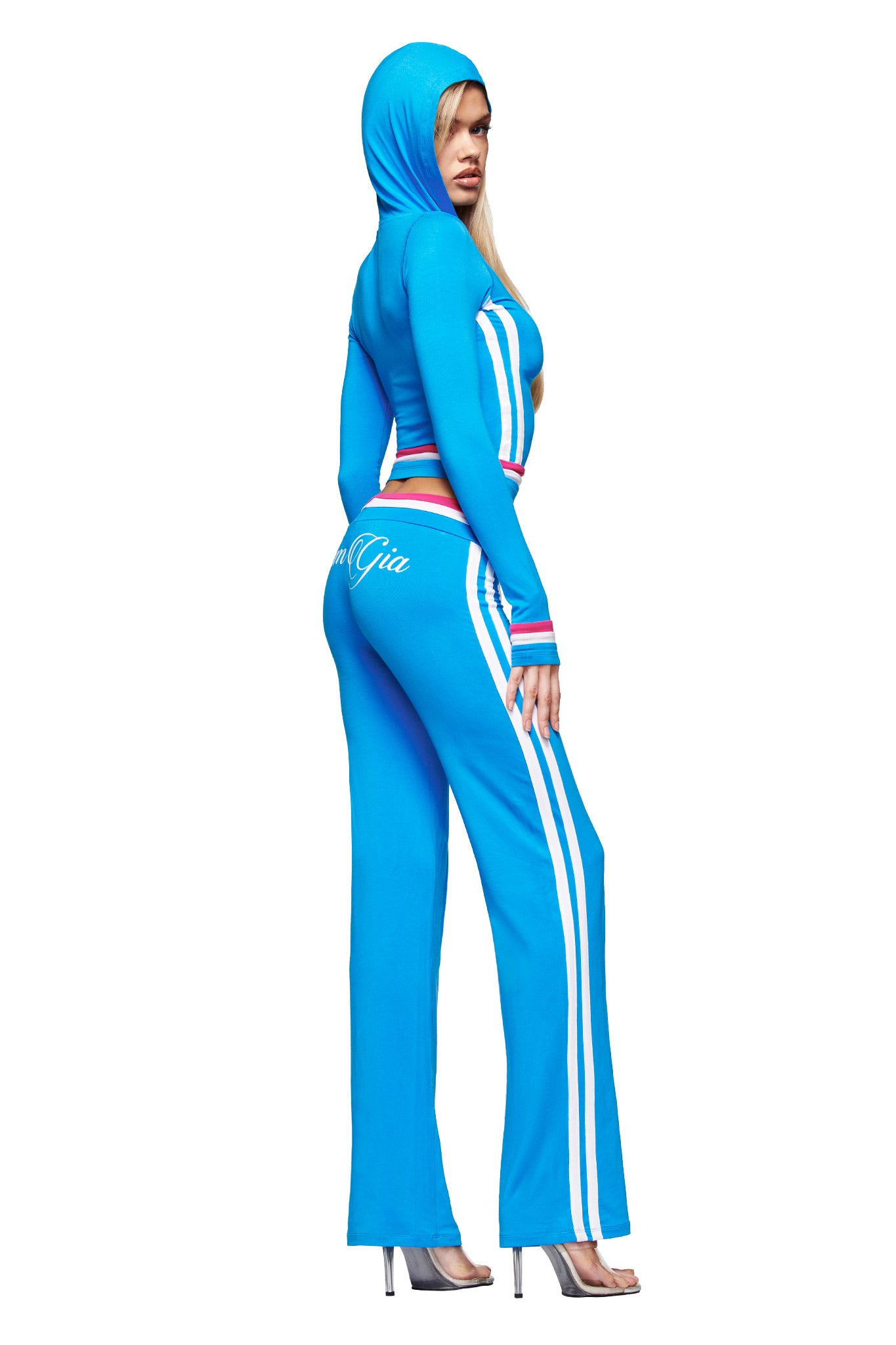 A woman poses sideways, looking over her shoulder, wearing the I.AM.GIA BLARE STRIPED HOODIE in sky blue with white and red stripes, paired with matching pants featuring white text and clear high-heeled shoes.