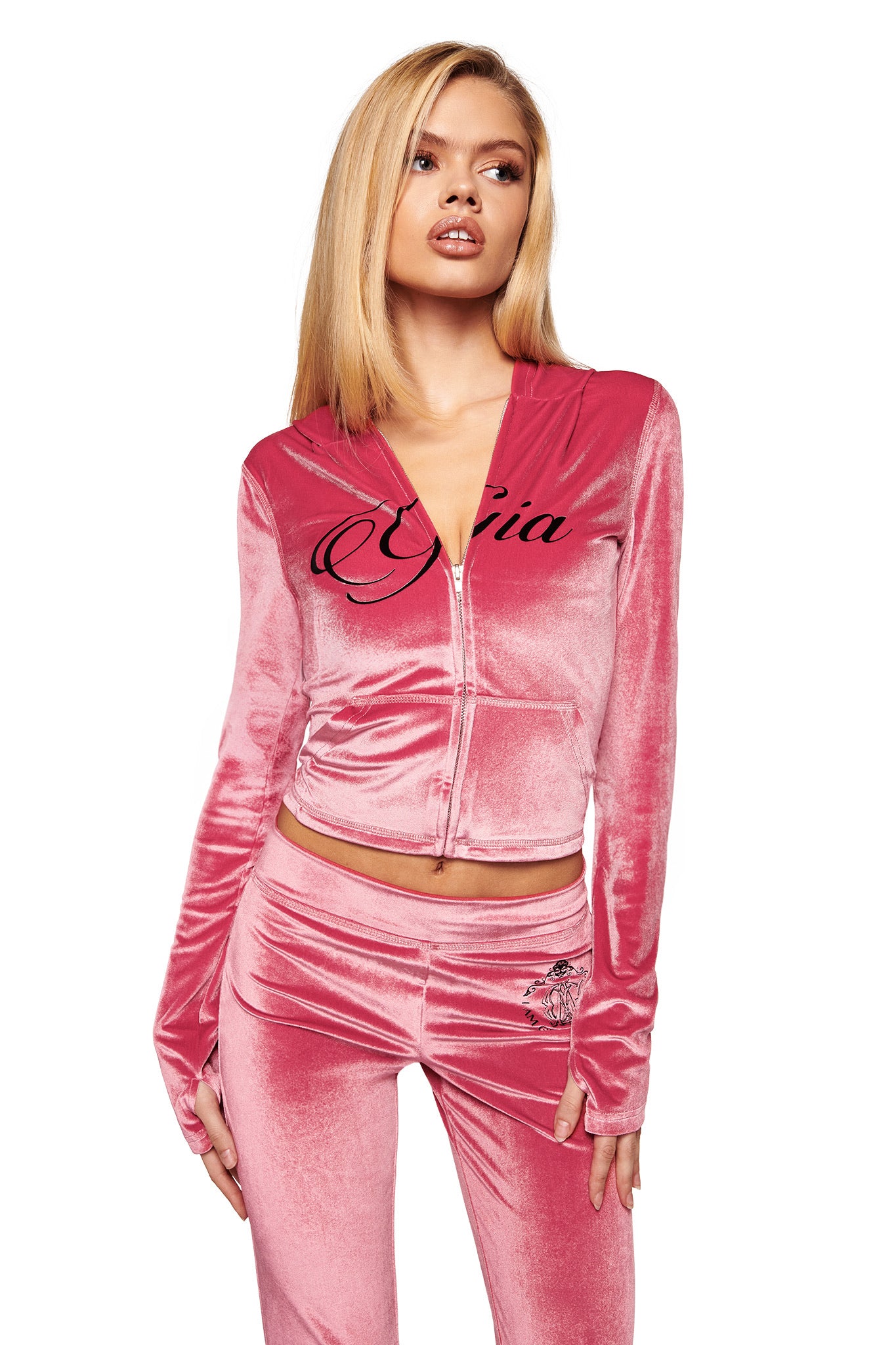 A woman with straight blonde hair models the I.AM.GIA BLARE VELOUR POCKET HOODIE in pink, featuring "Gia" in cursive and sparkling diamanté angel wings on the back, posing with her mouth slightly open and hand by her side.