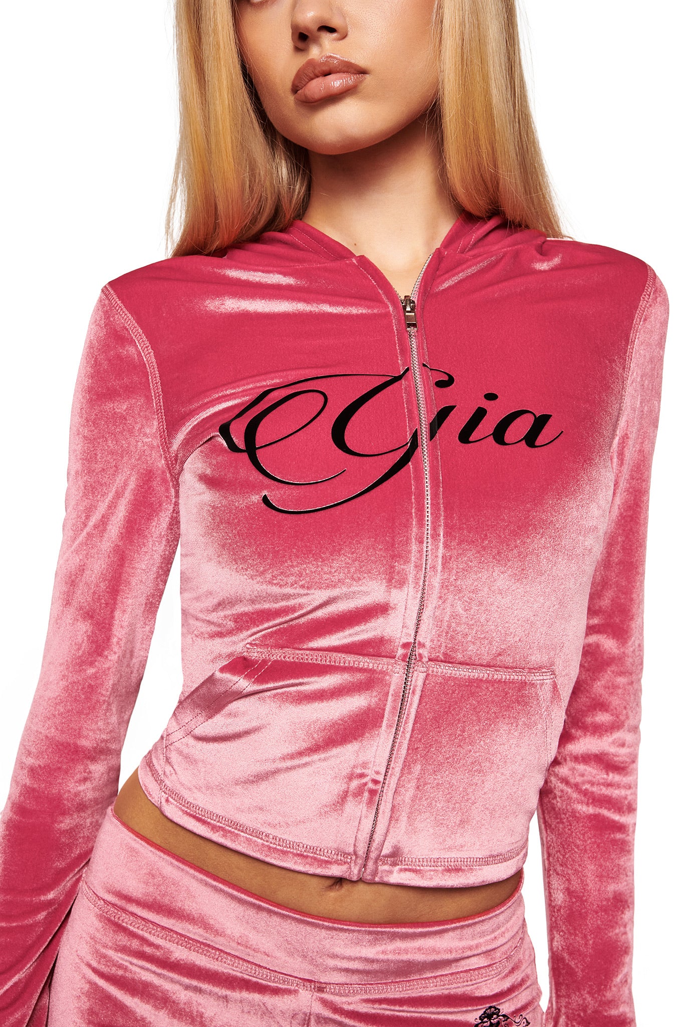 A woman with long blonde hair wears the I.AM.GIA BLARE VELOUR POCKET HOODIE in pink, featuring diamanté angel wings and "Gia" script on the front, paired with matching pink velour bottoms. Her face is partially out of frame.