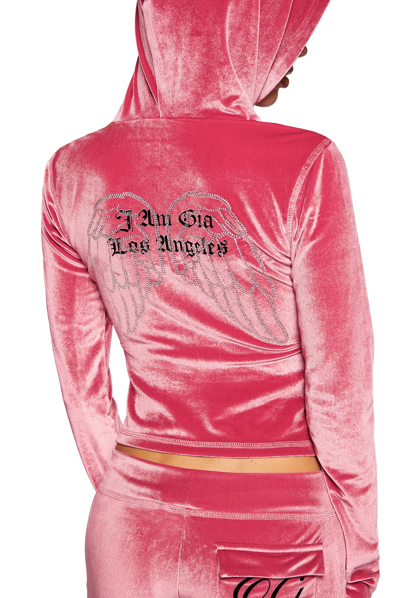 A person wears the I.AM.GIA BLARE VELOUR POCKET HOODIE in pink with diamanté angel wings and "I Am Gia Los Angeles" on the back. The hood is up, partly hiding their face and highlighting I.AM.GIA's signature vibe.