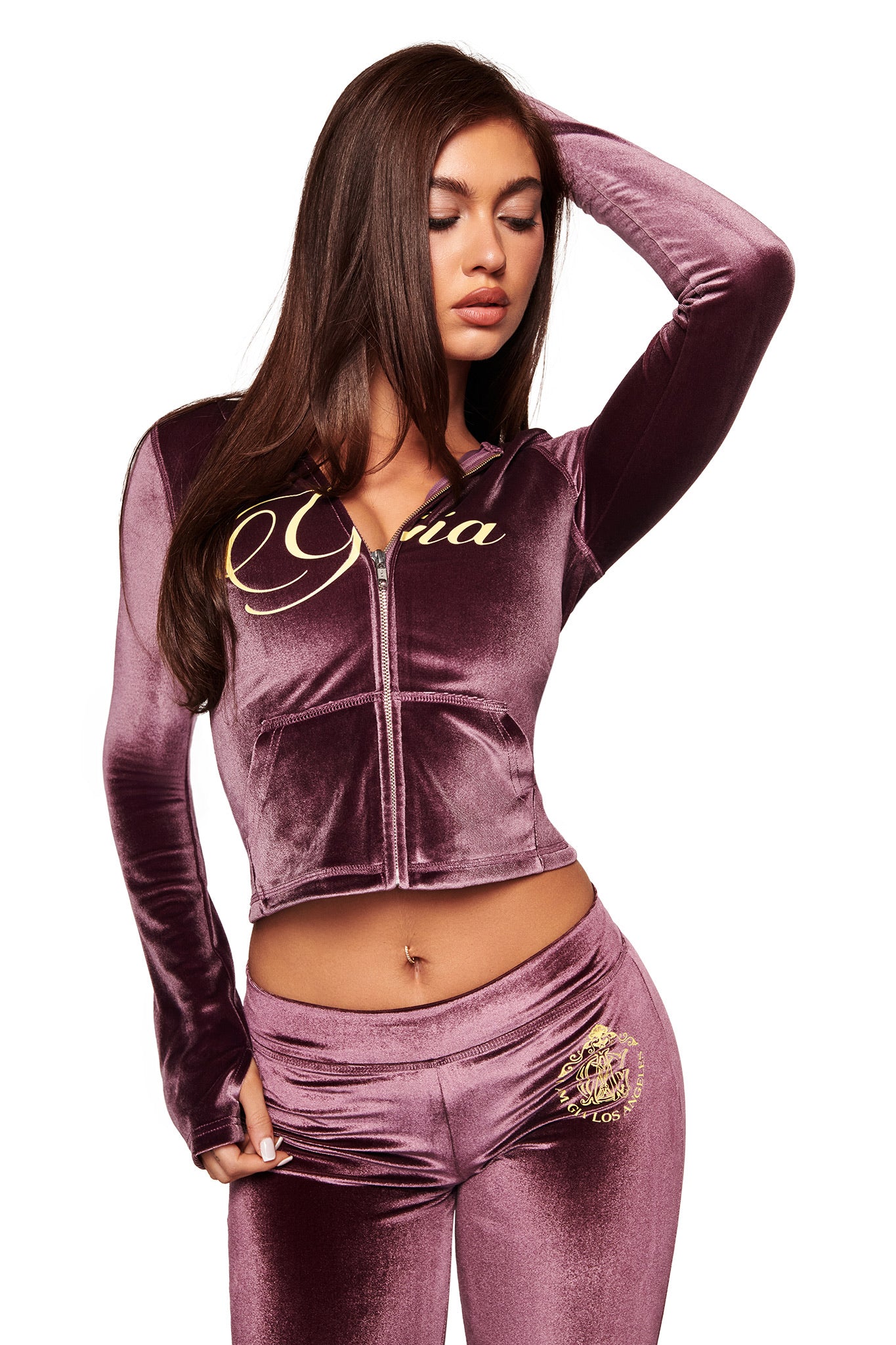 A woman wears the I.AM.GIA BLARE VELOUR POCKET TRACKPANT in purple with a matching zip-up jacket, posing with one arm raised and eyes closed against a white background.