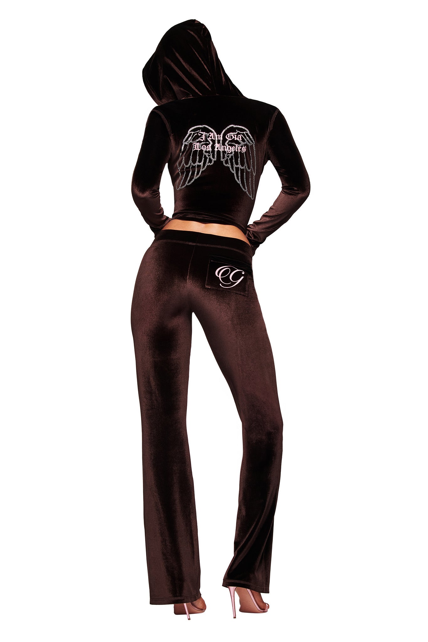 A person faces away, wearing the I.AM.GIA BLARE VELOUR POCKET HOODIE in chocolate with diamanté angel wings and GIA script logo on the back, paired with pants featuring decorative pockets and high heels.