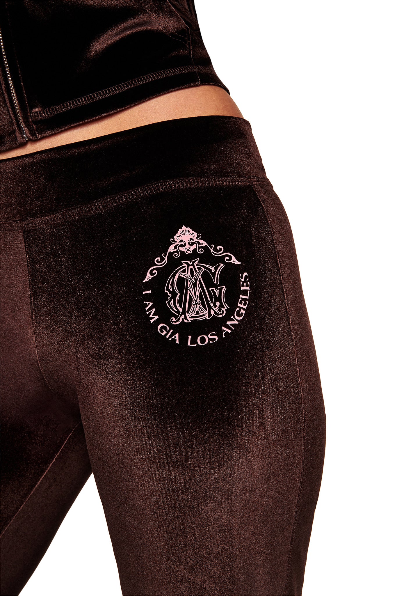 A close-up of someone wearing the I.AM.GIA BLARE VELOUR POCKET TRACKPANT in chocolate, featuring a sculpted fit, decorative crest, and “I.AM.GIA LOS ANGELES” in pink on the hip against a white background.