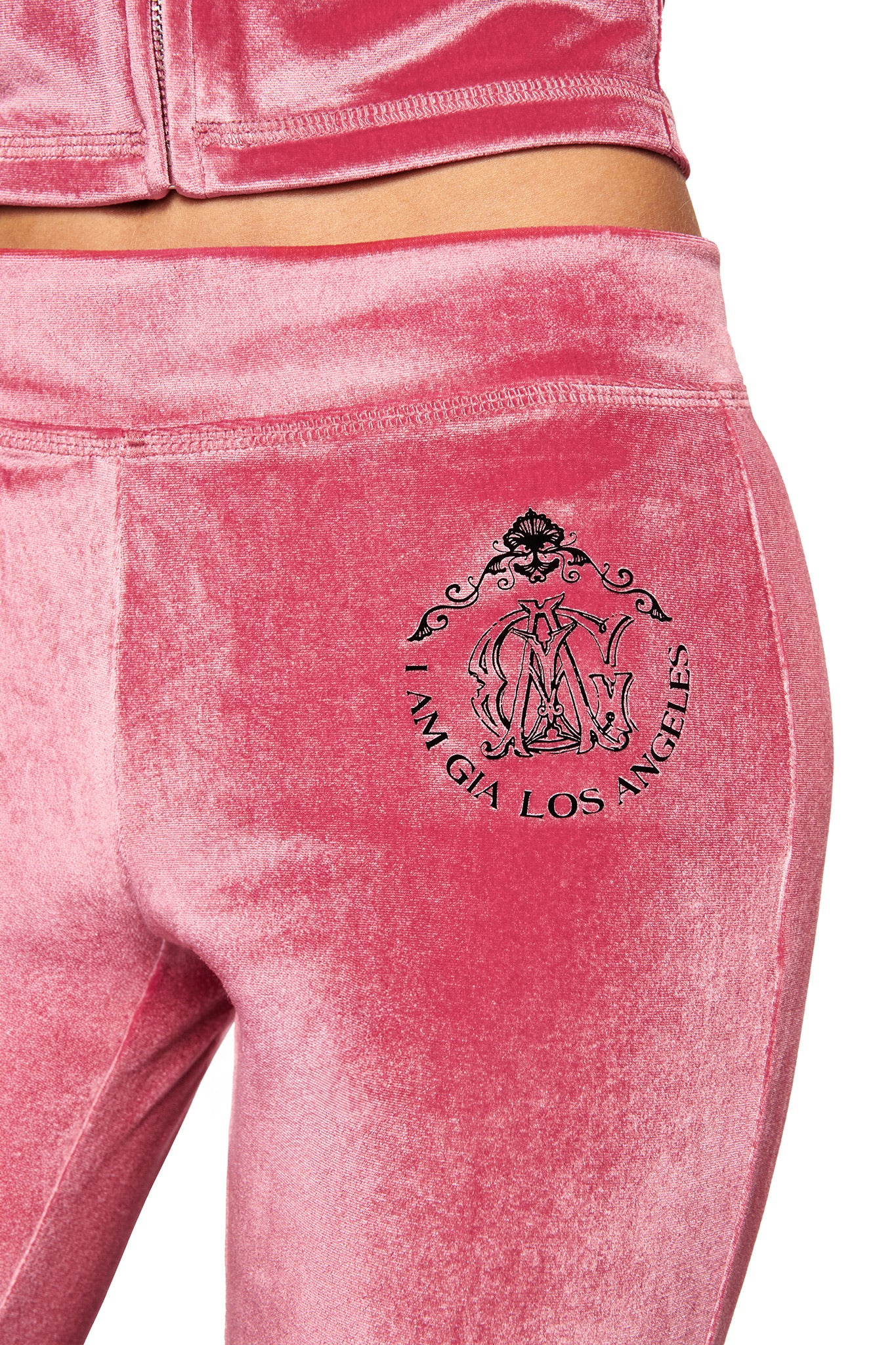 Close-up of a person wearing the I.AM.GIA BLARE VELOUR POCKET TRACKPANT in pink, featuring a mid rise waist and an embroidered black crest with GIA script logo on the upper hip. Matching pink jacket is partially visible.