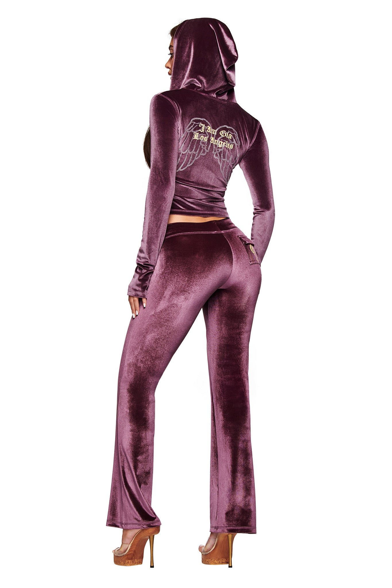 A woman faces away in the BLARE VELOUR POCKET HOODIE - PURPLE by I.AM.GIA, featuring embroidered angel wings and the GIA logo on the back, styled with high-heeled platform shoes.