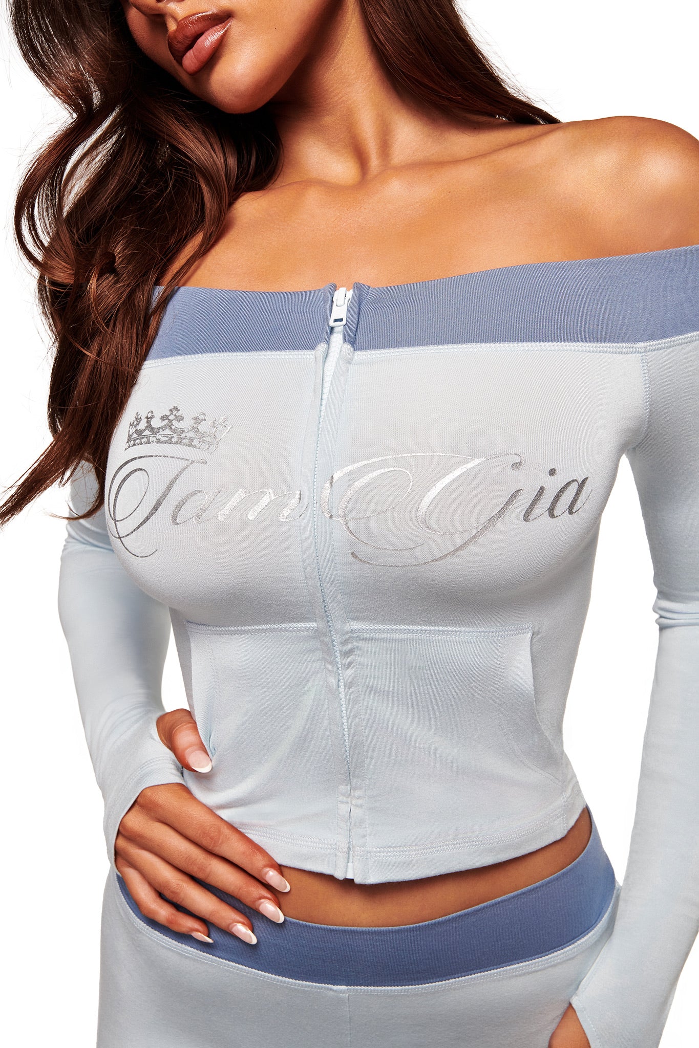 A woman models the I.AM.GIA BLARE OFF SHOULDER TOP - TWO TONE POWDER BLUE, a zip-up, buttery-soft viscose top featuring "Tam Gia" and a crown graphic on the chest, paired with matching pants, one hand on her waist.