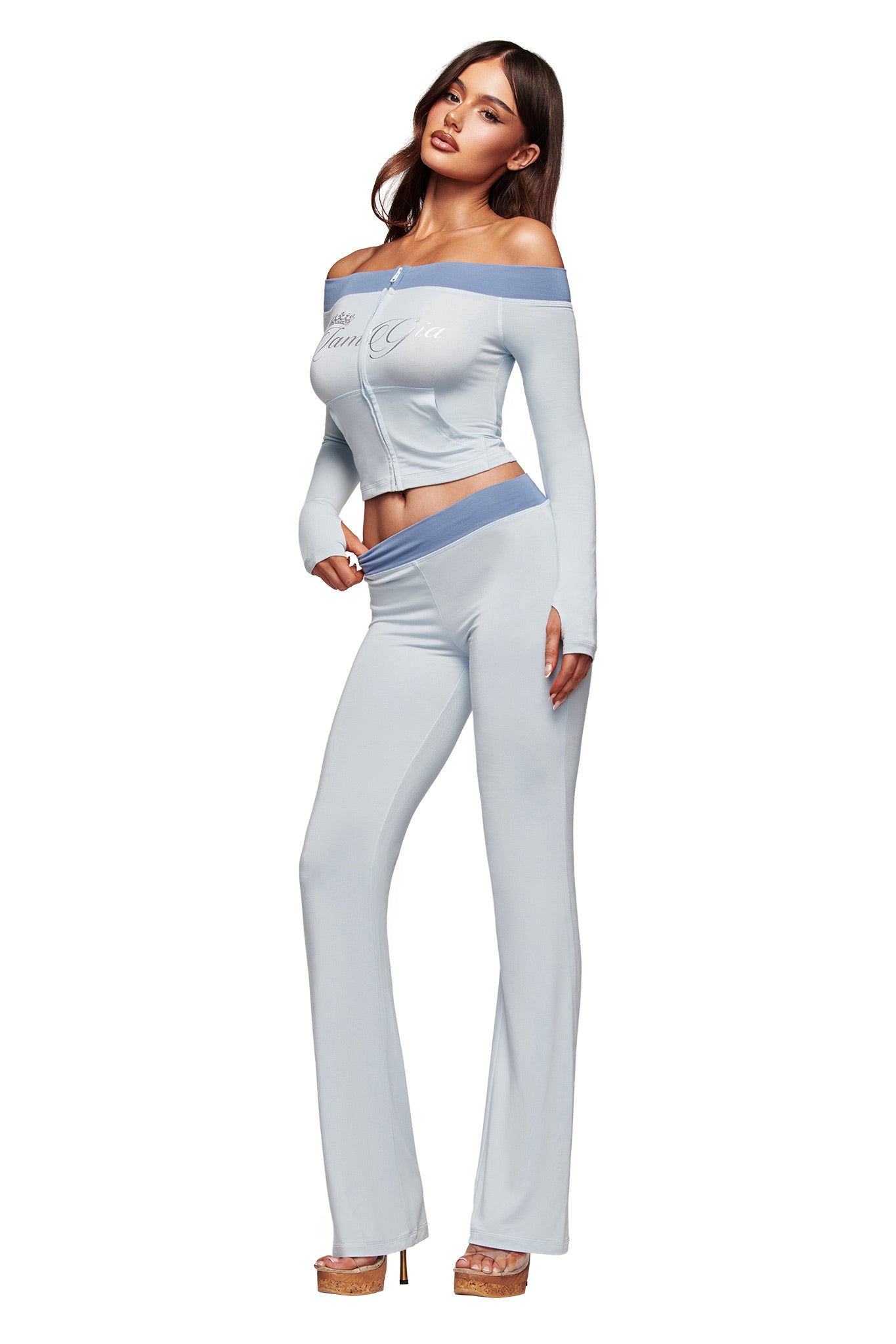 A woman models the I.AM.GIA BLARE OFF SHOULDER TOP - TWO TONE POWDER BLUE, a buttery-soft viscose light blue zip-up top with matching high-waisted flared pants, tan heels, and blue accents against a white background.