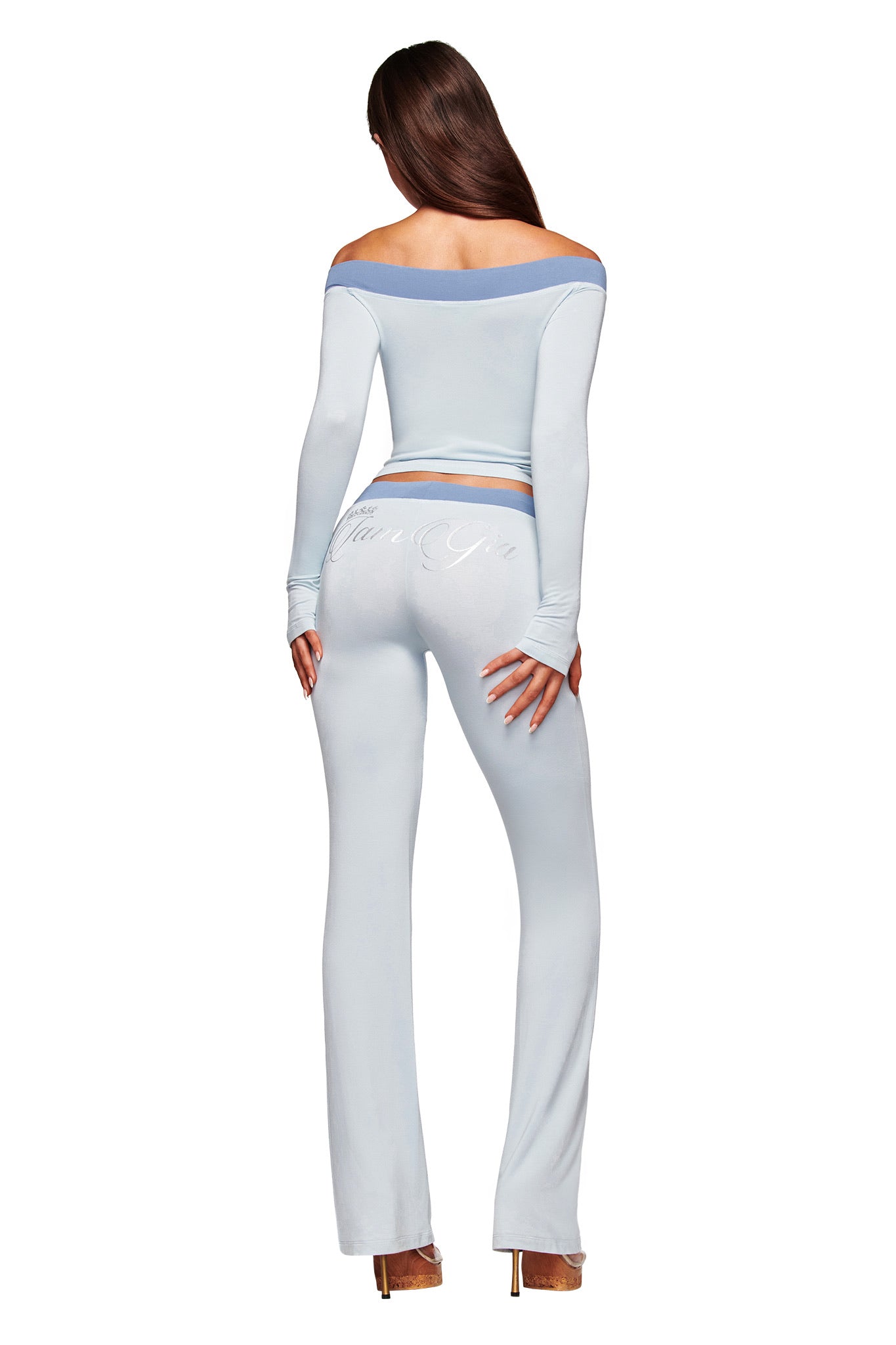 A woman with long brown hair faces away, wearing the I.AM.GIA BLARE OFF SHOULDER TOP in two tone powder blue and matching fitted pants, made from buttery-soft viscose, plus gold heels, against a white background.