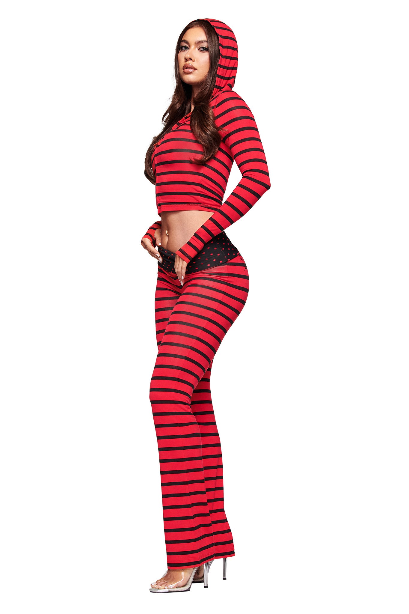 A woman wears the I.AM.GIA BLARE FOLDOVER TRACKPANT in red and black stripe with a matching hooded crop top, black studded belt, and clear heels, posing against a white background.