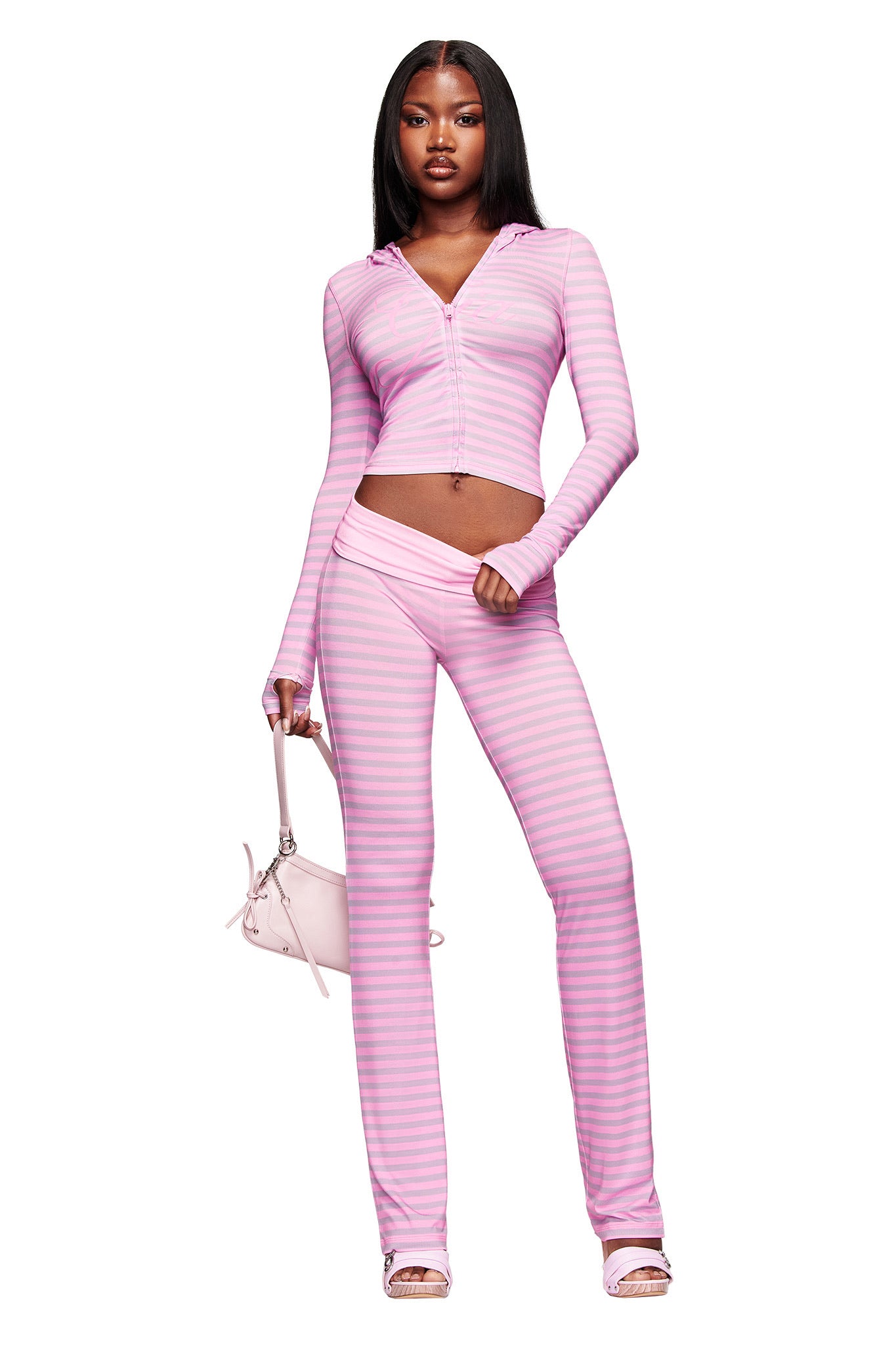A woman confidently wears the I.AM.GIA BLARE HOODIE - LIGHT PINK STRIPE as a long-sleeve crop top with matching flared pants, paired with a pink handbag and white heeled sandals against a plain white background.