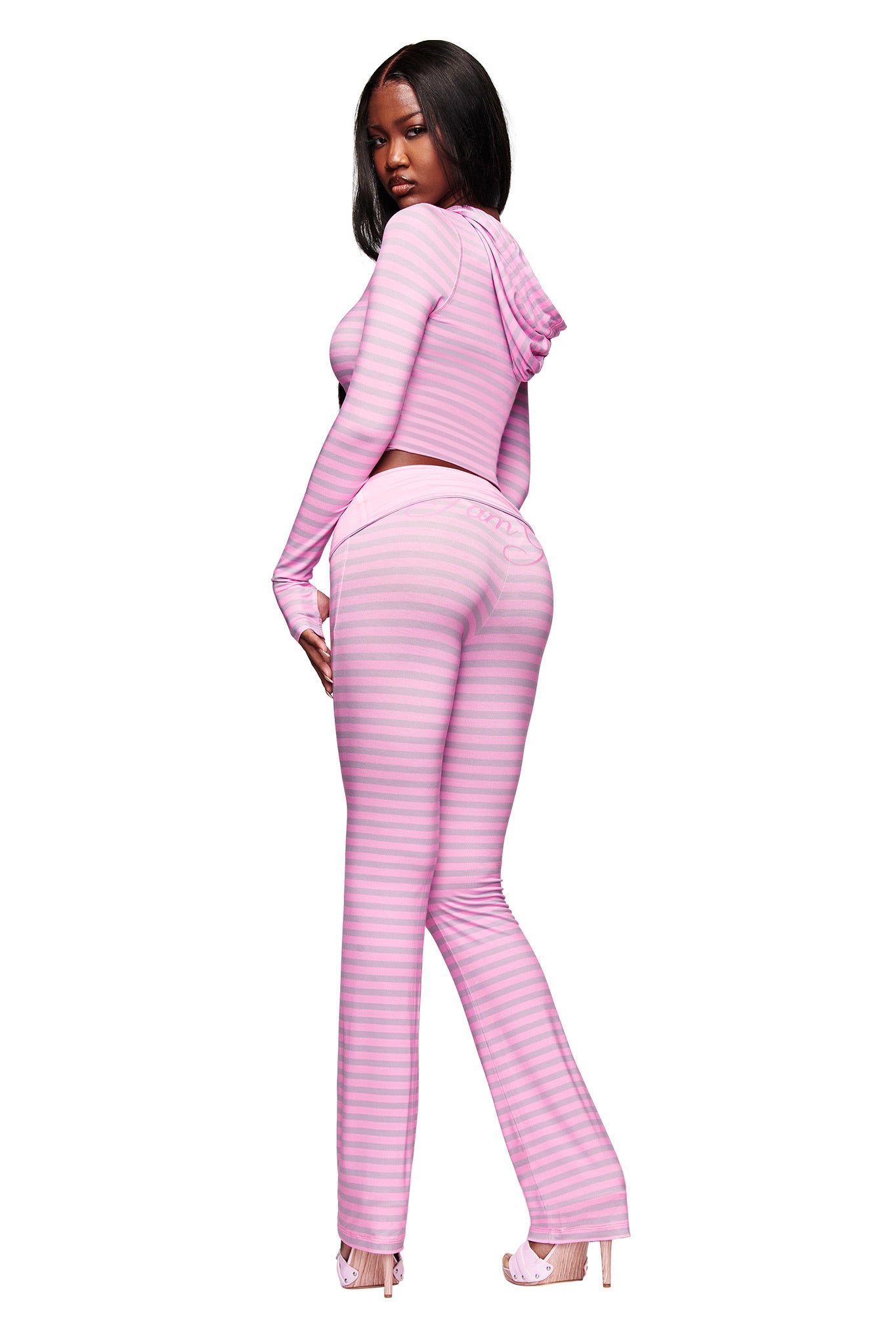 A woman looks over her shoulder, wearing the I.AM.GIA BLARE FOLDOVER TRACKPANT in Light Pink Stripe with a matching pink and light purple striped hooded crop top. The background is white.