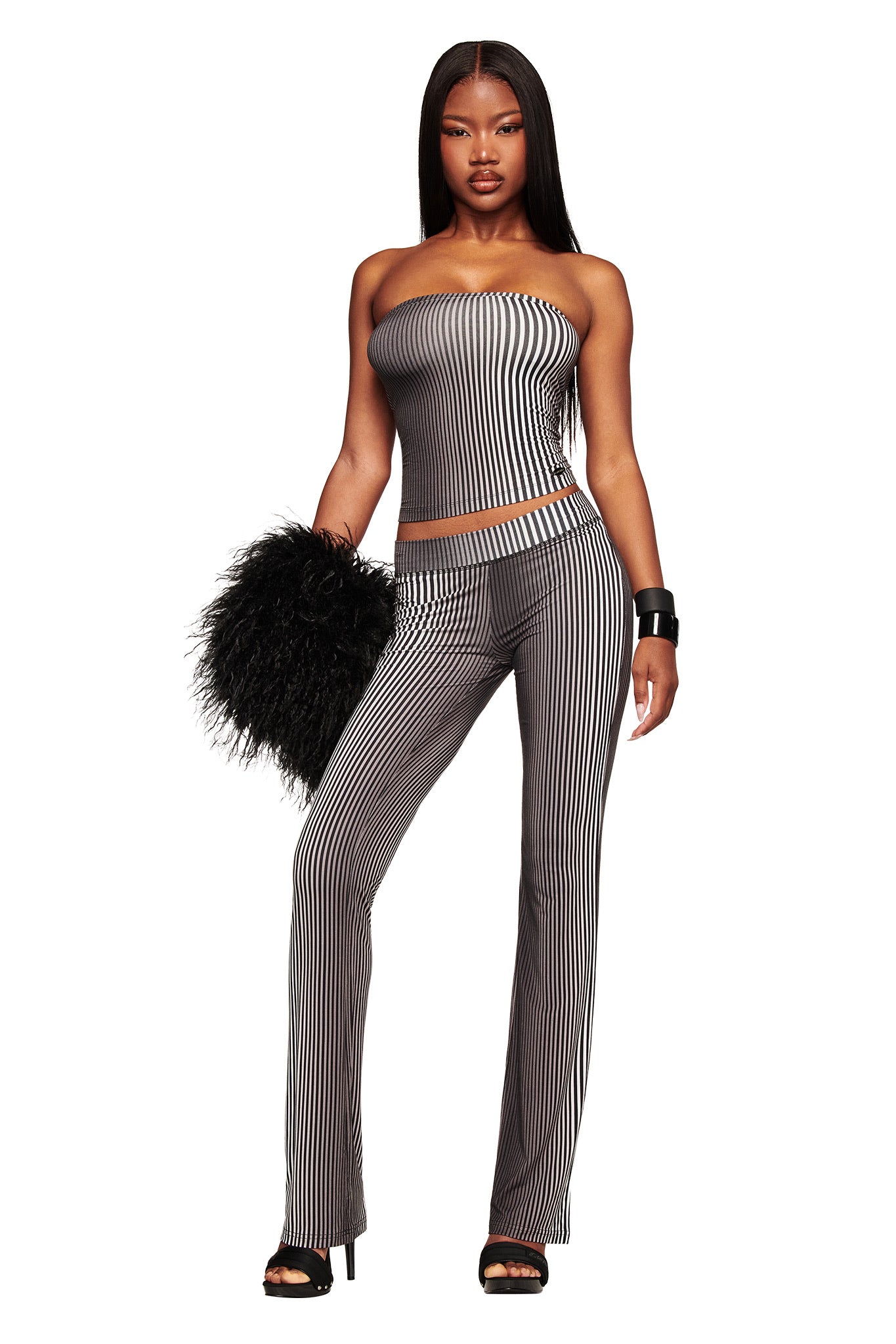 A woman stands confidently in the I.AM.GIA TRINITY 2-IN-1 SKIRT PANT - STRIPE, paired with a stylish top, black feathered clutch, black heels, and a chunky bracelet.