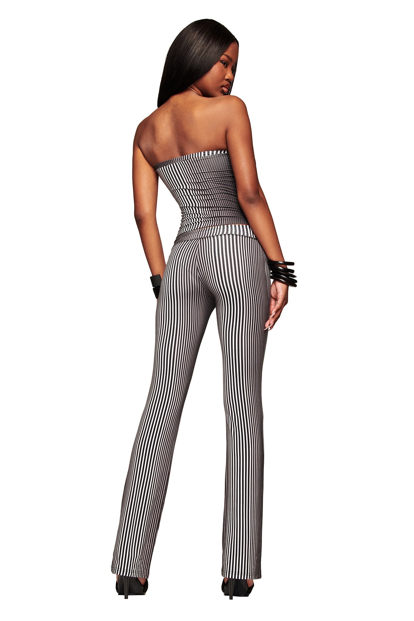 A woman faces away, wearing the I.AM.GIA TRINITY 2-IN-1 SKIRT PANT - STRIPE with black heels, straight long hair, and black bangles on one wrist, set against a white background.