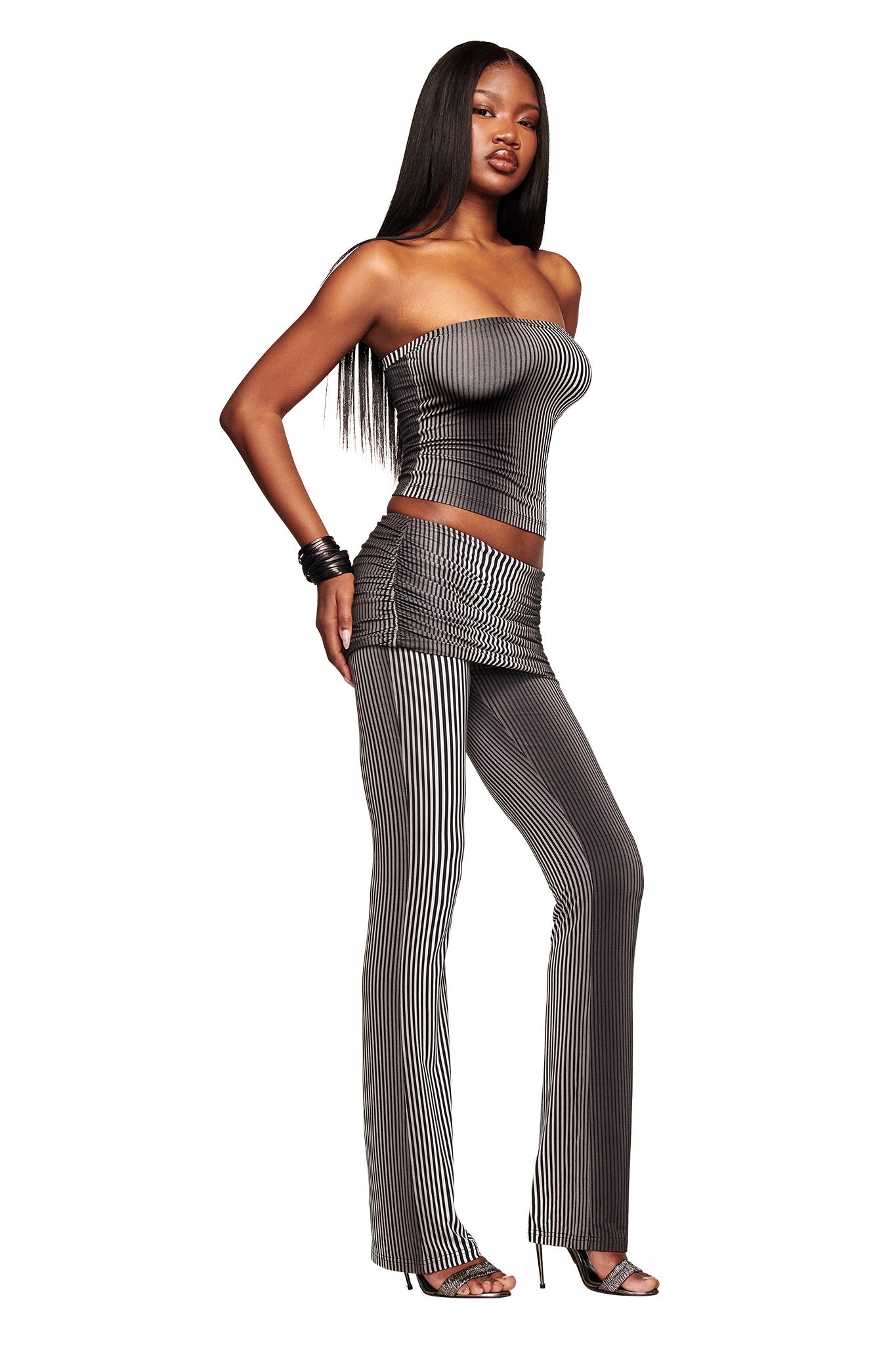 A woman with long straight hair wears I.AM.GIA’s TRINITY 2-IN-1 SKIRT PANT - STRIPE and a strapless bamboo viscose jersey top, styled with hoop earrings, bangles, and high heels against a white background.