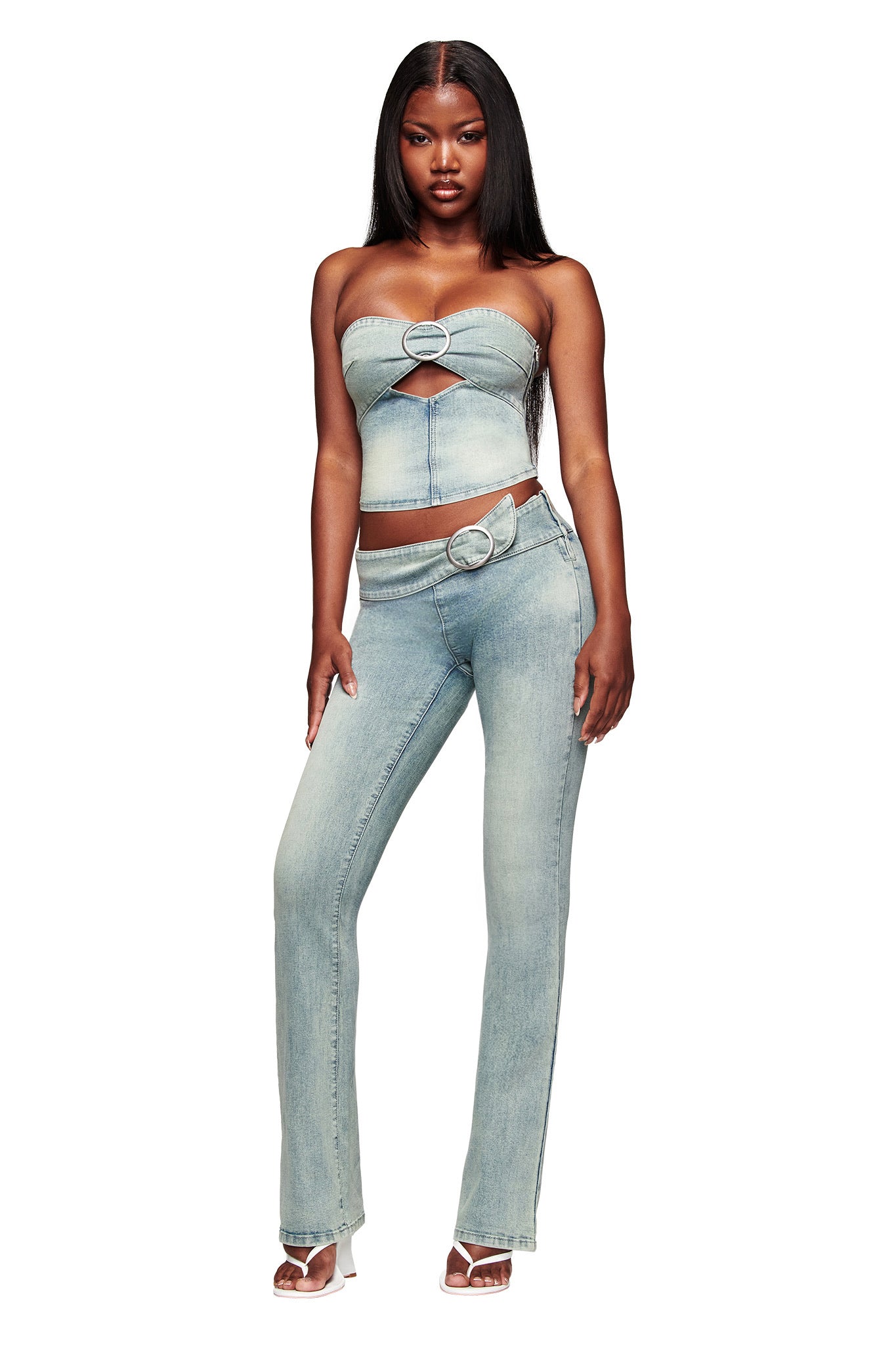 A woman poses in the I.AM.GIA JENN JEAN - BLUE set, featuring a strapless denim top with cutout and metal details, paired with matching mid-rise flared pants and white heels, against a white background.