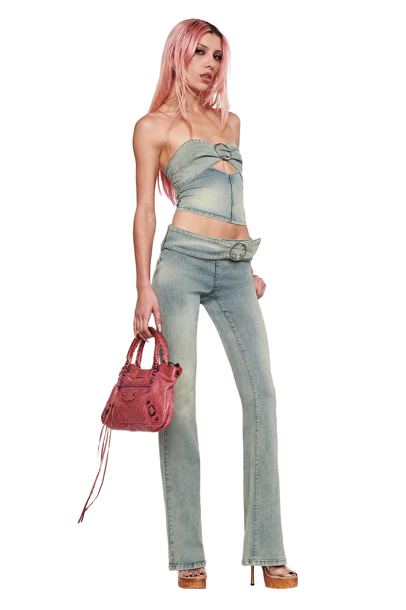 A woman with long pink hair poses confidently against a white background, wearing the I.AM.GIA JENN JEAN - BLUE flared denim pants with a strapless top, wide belt, platform heels, and holding a pink handbag.