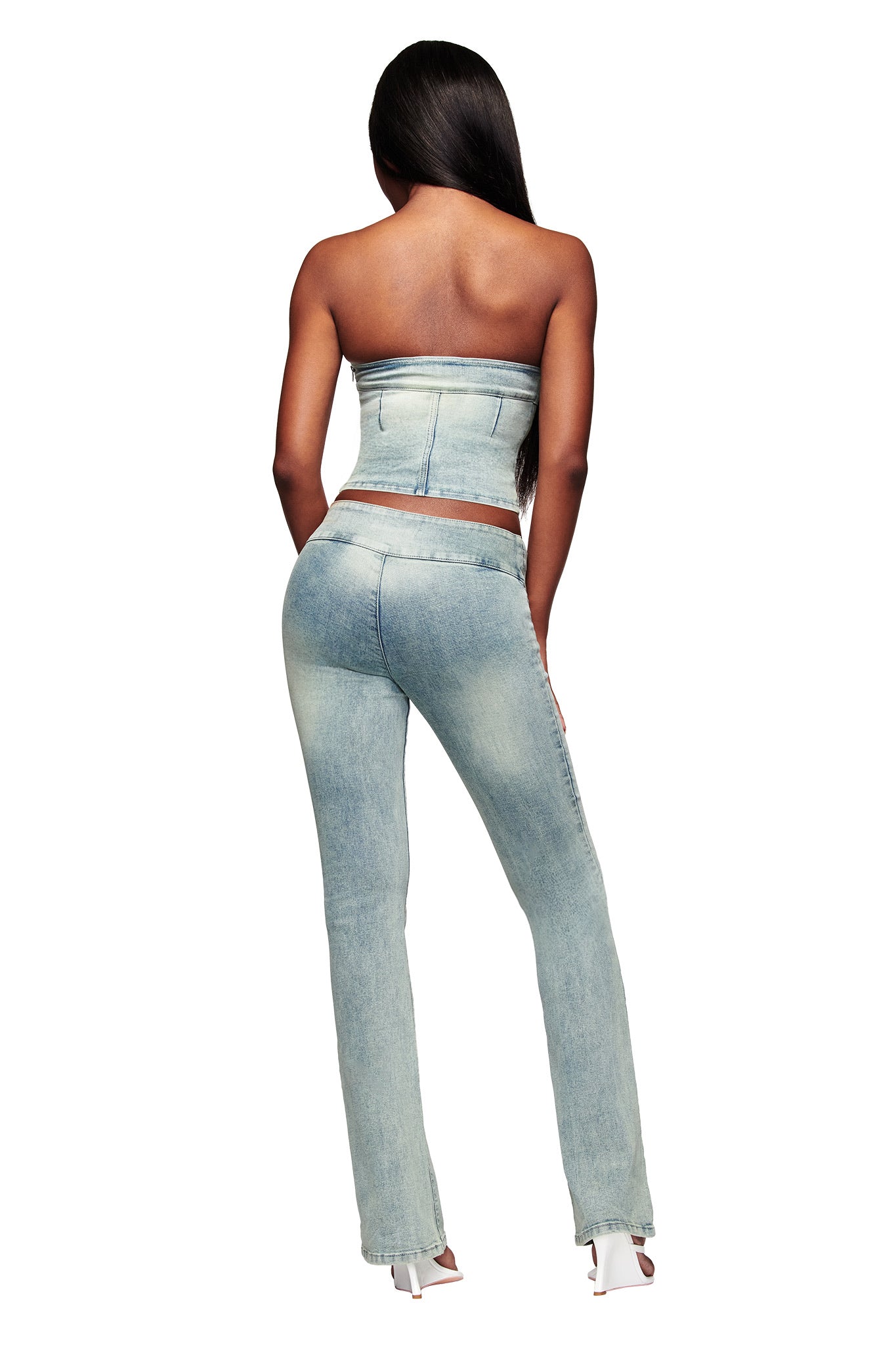 A woman with long dark hair wears I.AM.GIA's JENN JEAN - BLUE, featuring light wash mid-rise flared denim pants and a matching strapless top. She pairs the look with white heels against a white background.