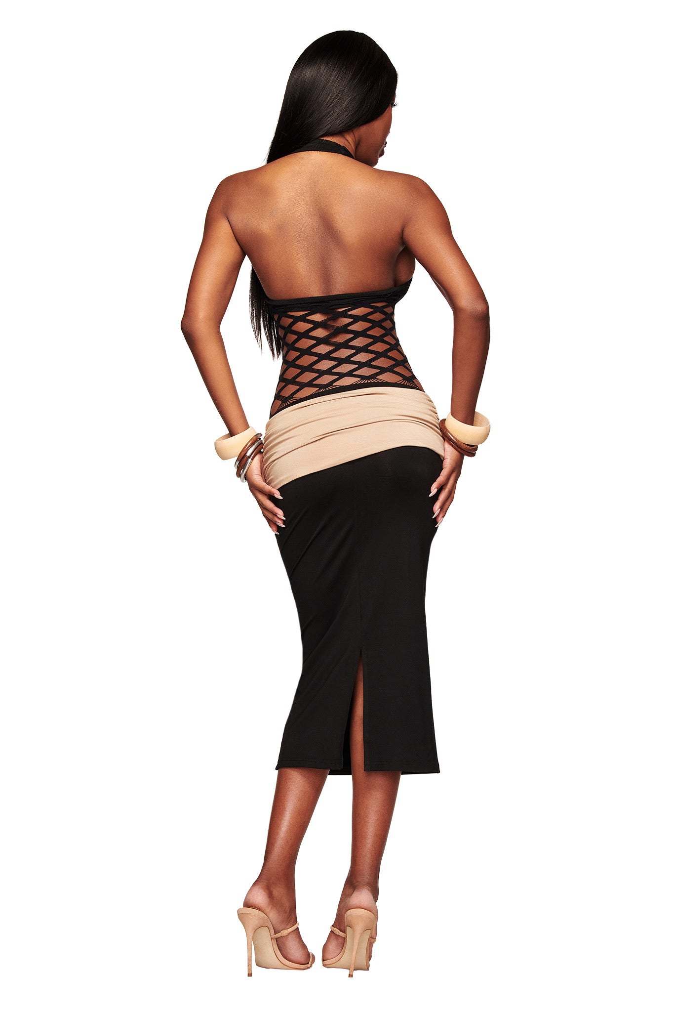 A woman with long dark hair stands with her back to the camera, wearing the I.AM.GIA ZOREEN MIDI DRESS in black, featuring a fishnet bodice panel and crisscross mesh details. She pairs it with high heels and chunky bracelets against a white background.