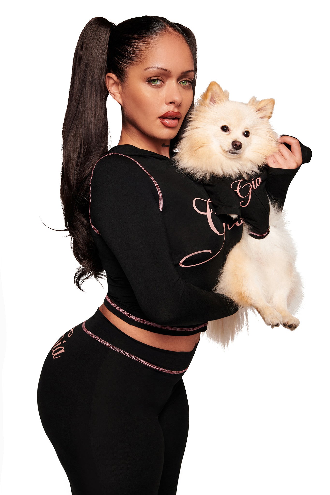A woman with long dark hair in high pigtails wears black activewear with pink trim, holding a fluffy white Pomeranian in the I.AM.GIA BLARE BEST FRIEND DOG HOODIE - BLACK. Both face the camera against a crisp white background.
