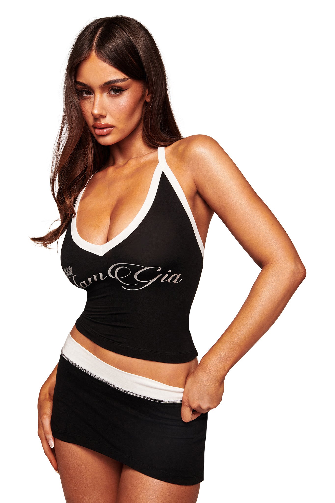 A woman with long brown hair wears a black halter top with white trim and "I.AM.GIA" branding, paired with the BLARE MICRO SKORT in black and white from I.AM.GIA, featuring a mid-rise waist, posing against a white background.
