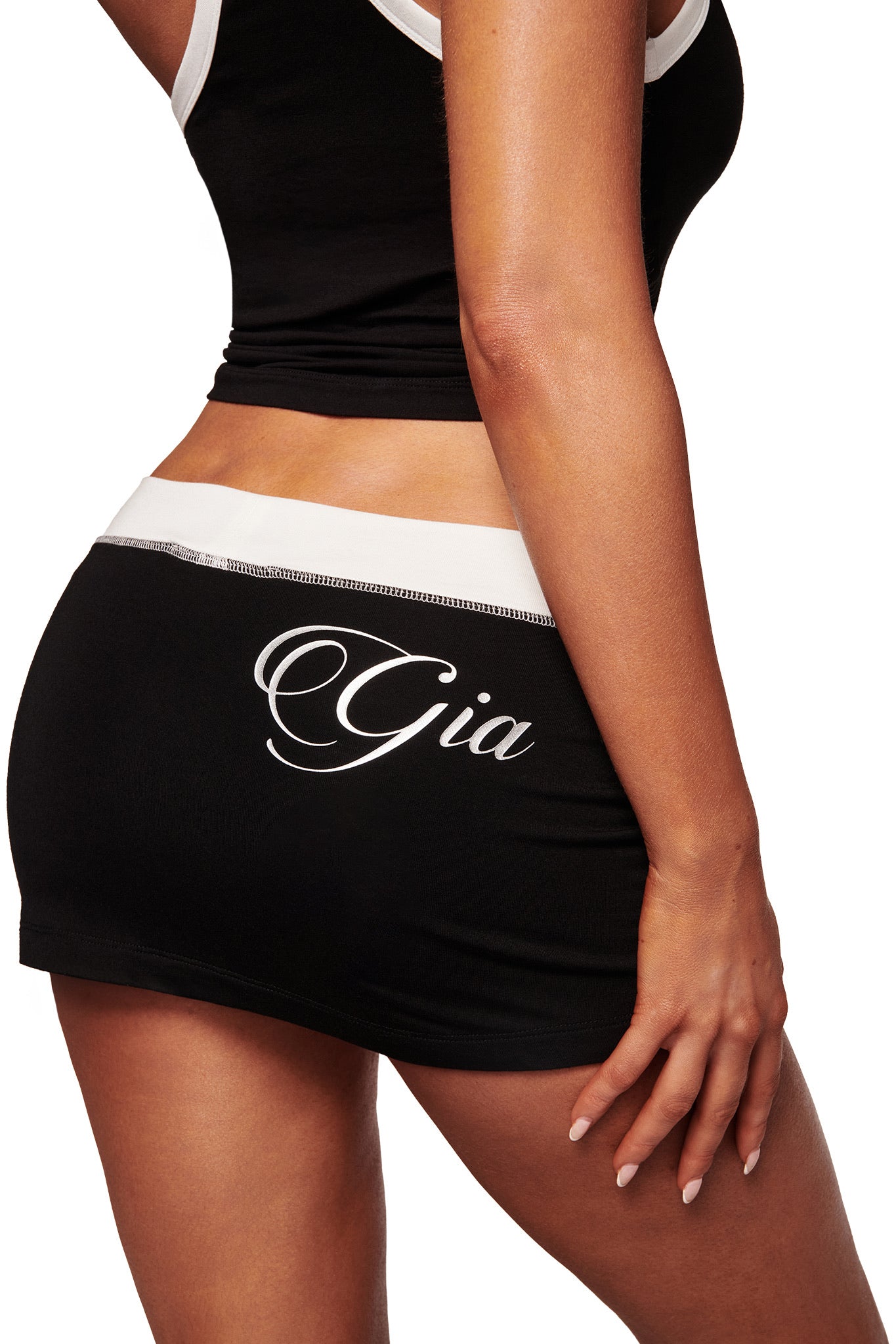 A woman wears the I.AM.GIA BLARE MICRO SKORT in black and white with a matching tank, both crafted from buttery-soft viscose. "Gia" script adorns the back; only her torso and upper legs are shown as her hand rests on her hip.