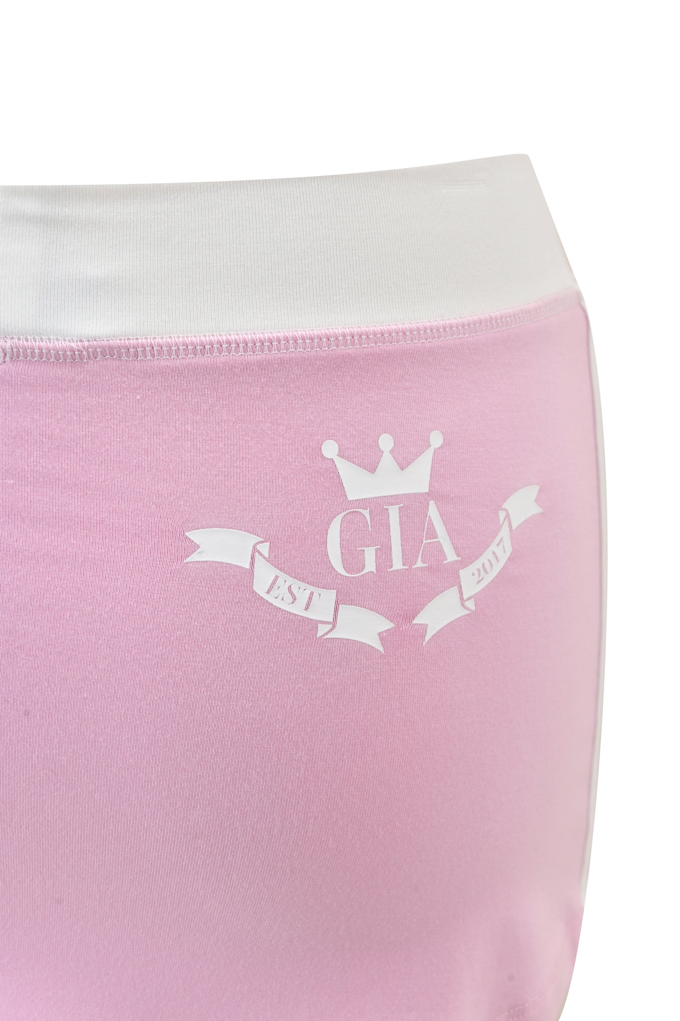 A close-up of the I.AM.GIA BLARE PIPING TRACKPANT in baby pink and white, showing buttery-soft viscose fabric, a white waistband, a crown graphic above "GIA," and a ribbon banner with "EST 2017.