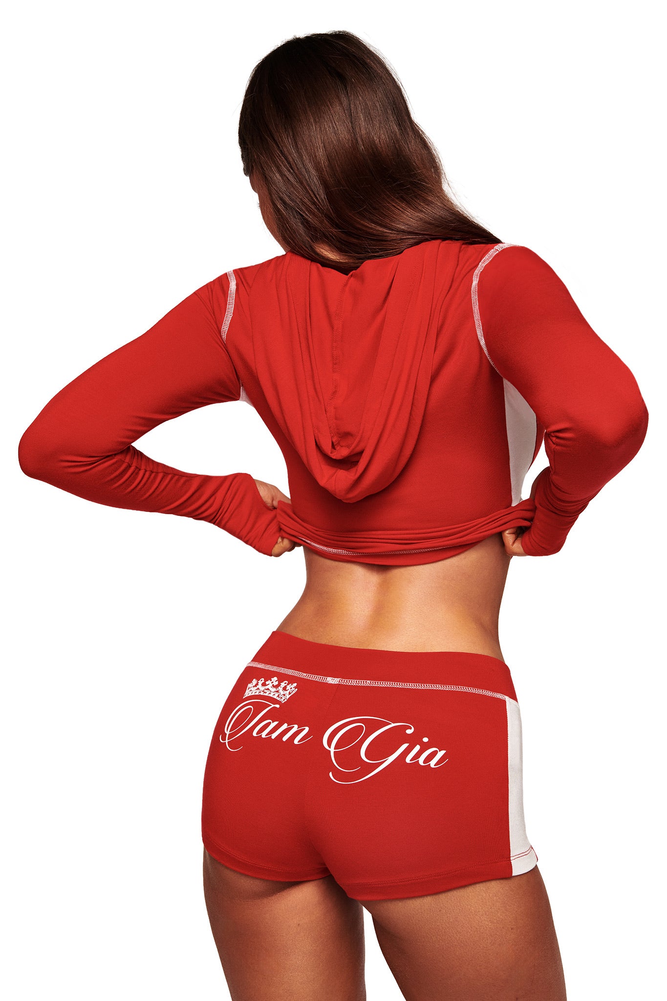 A woman with long brown hair faces away, wearing the I.AM.GIA BLARE PANELLED SHORT in red with white trim and a matching red hooded crop top. "I.AM.GIA" in bold white script decorates the back of the sporty bamboo-fabric shorts.