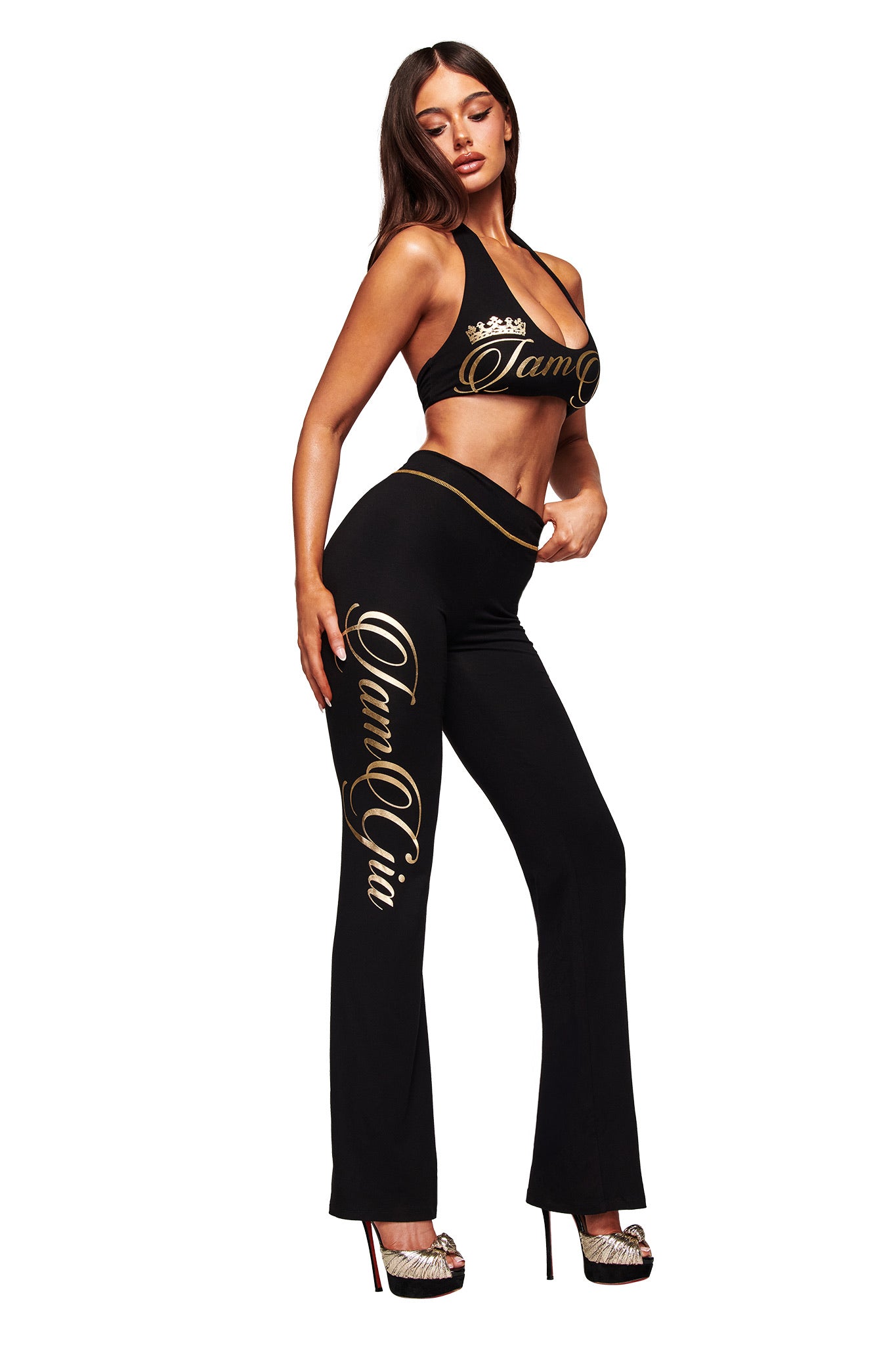 A woman models the I.AM.GIA BLARE FORM HALTER CROP TOP in black and gold, featuring bold gold branding, paired with matching fitted pants. She completes the look with high heels against a white background.