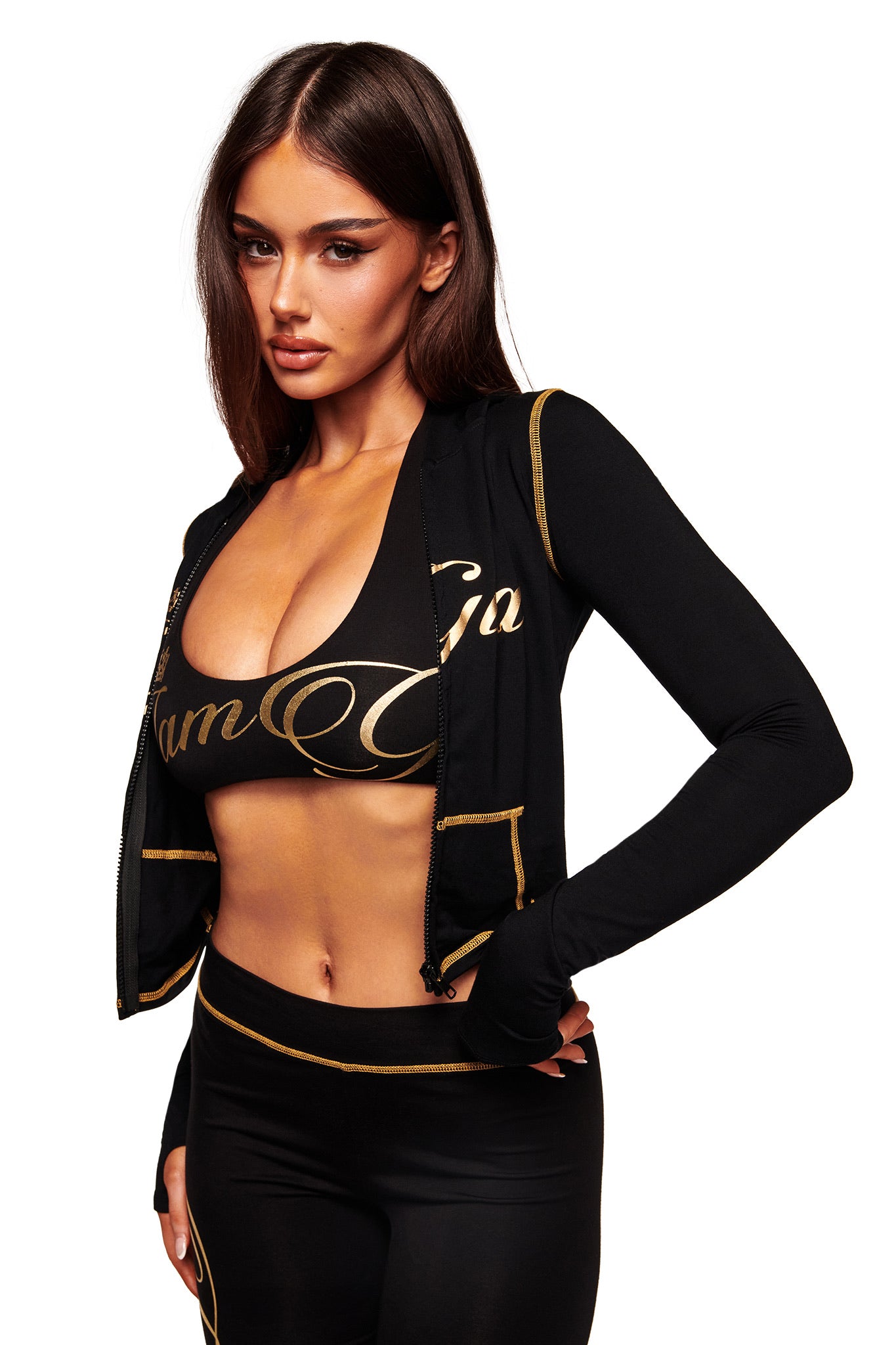 A woman models the I.AM.GIA BLARE FORM MULTI POCKET HOODIE in black and gold, paired with a matching crop top and high-waisted leggings. She poses against a white background with her long brown hair down, gazing to the side.