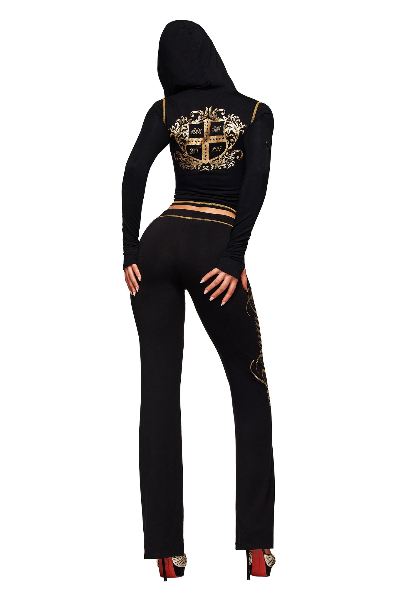 A person wearing the I.AM.GIA BLARE FORM MULTI POCKET HOODIE in black and gold stands with their back to the camera, hands at their sides, paired with red high heels.